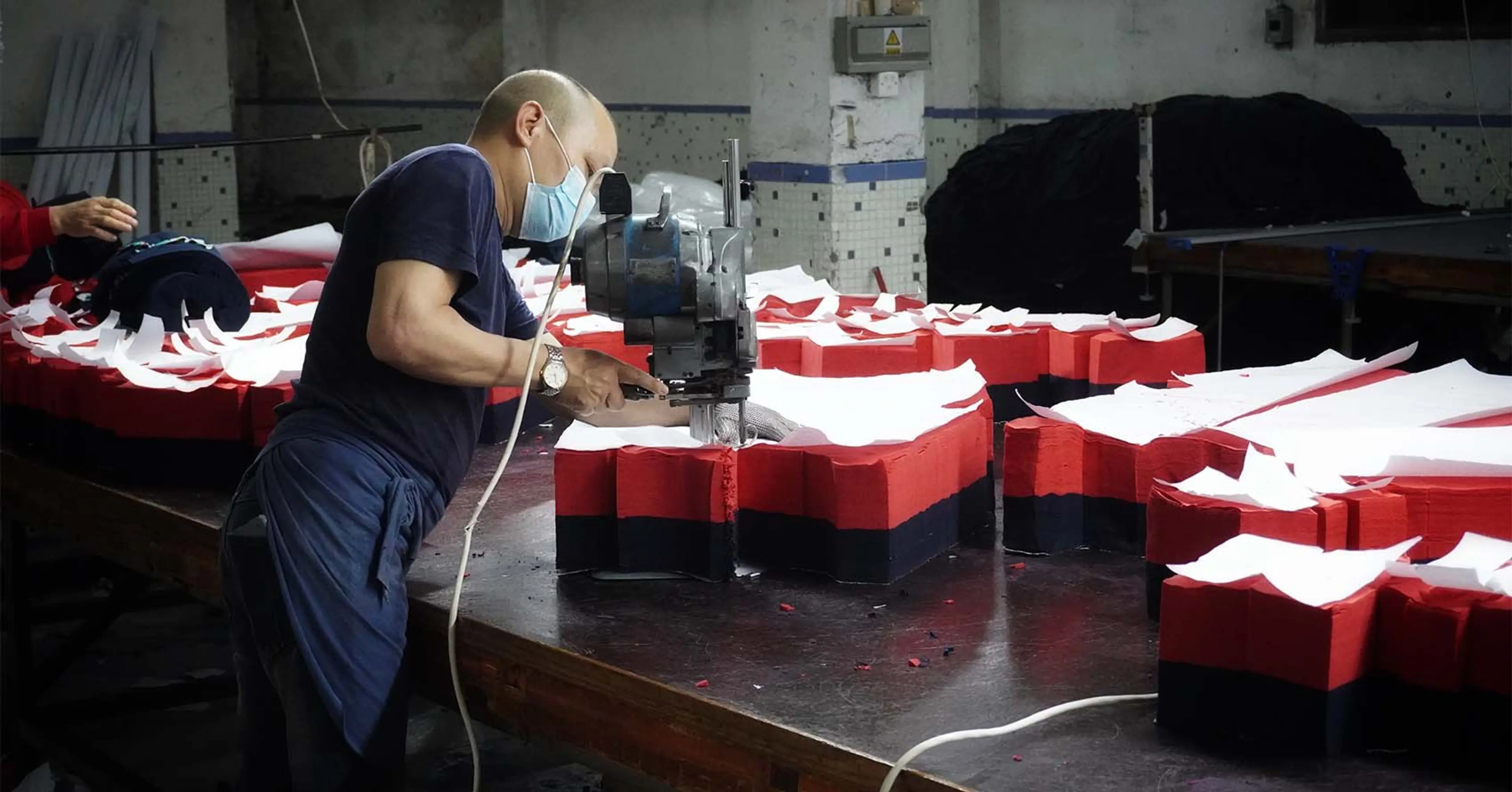 En person med munskydd arbetar med en maskin för att skära eller forma skumgummi i röd, svart och vit färg på ett stort arbetsbord. Industrimiljö med fokus på hantverksmässigt detaljarbete.