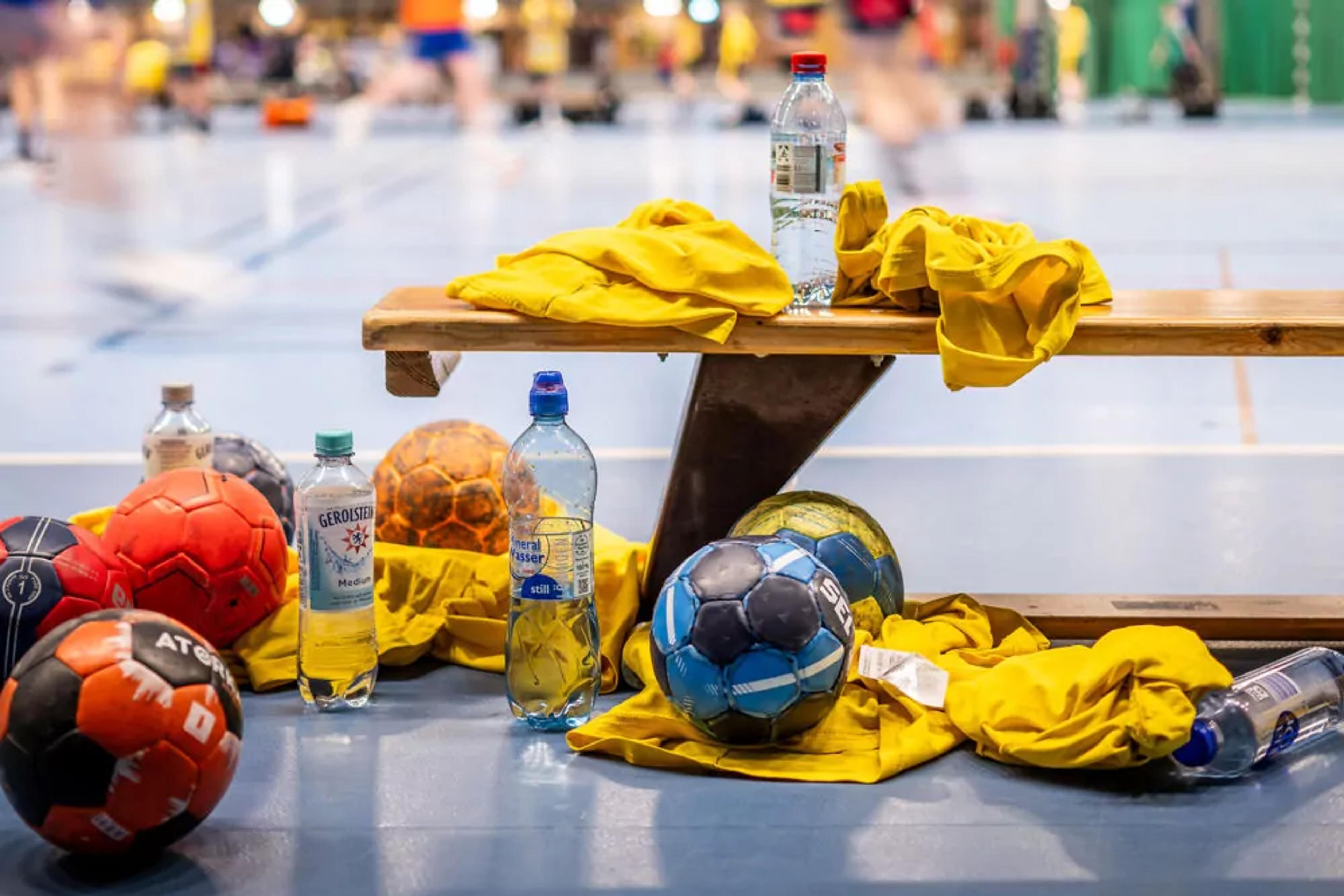 Flera handbollar i olika färger ligger på golvet tillsammans med gula träningsvästar och vattenflaskor vid en bänk i en sporthall. I bakgrunden syns suddiga rörelser från personer i aktivitet.