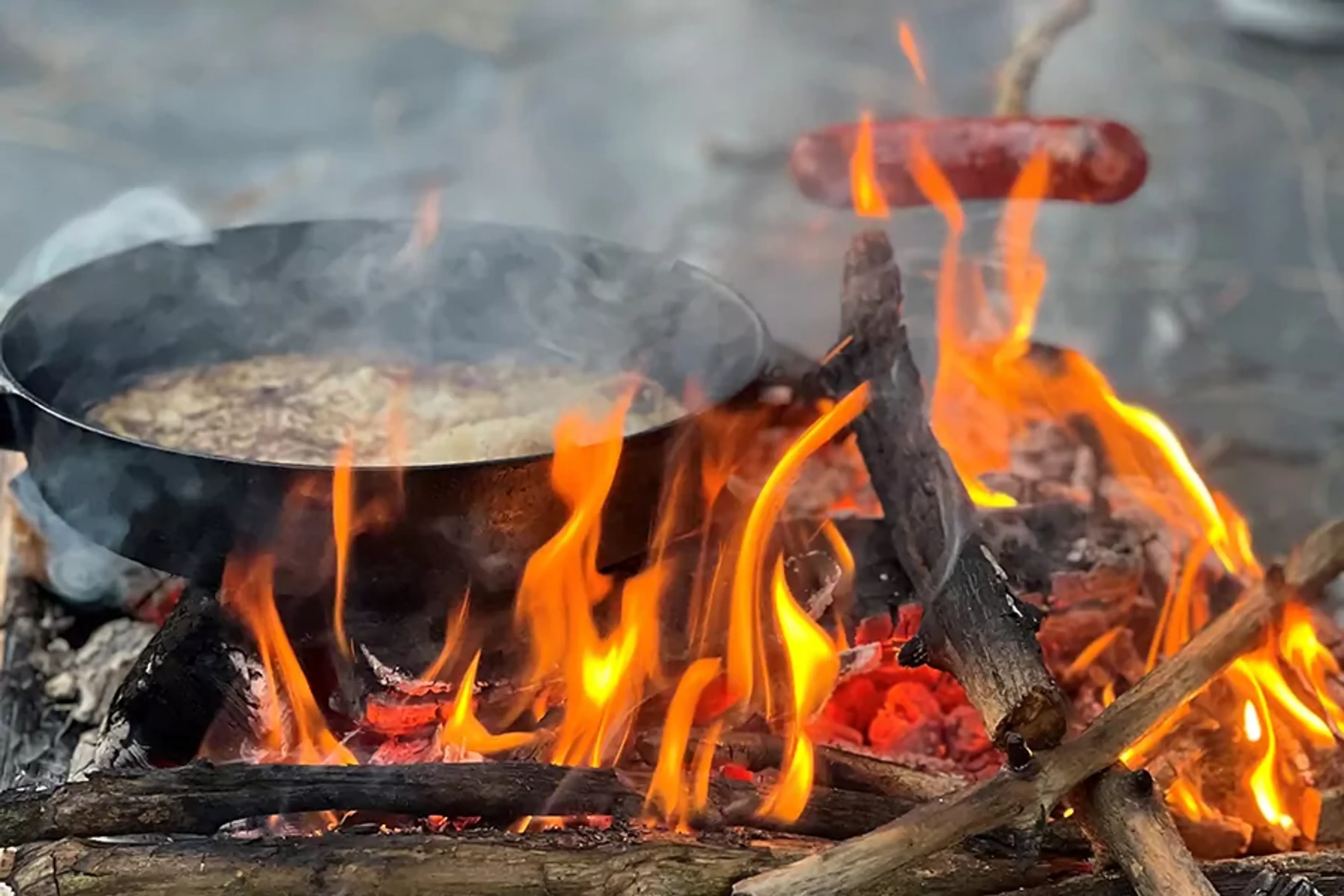 En gryta står på öppen eld med lågor som dansar runt vedträn, samtidigt rostas en korv på en pinne över elden. Rök och värme skapar en äkta utomhuskänsla.