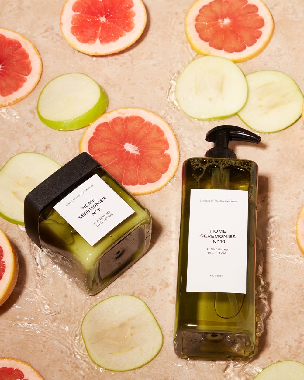 En kroppslotion och en duschtvål i gröna transparenta förpackningar, omgivna av skivor av grapefrukt och äpple i vatten.