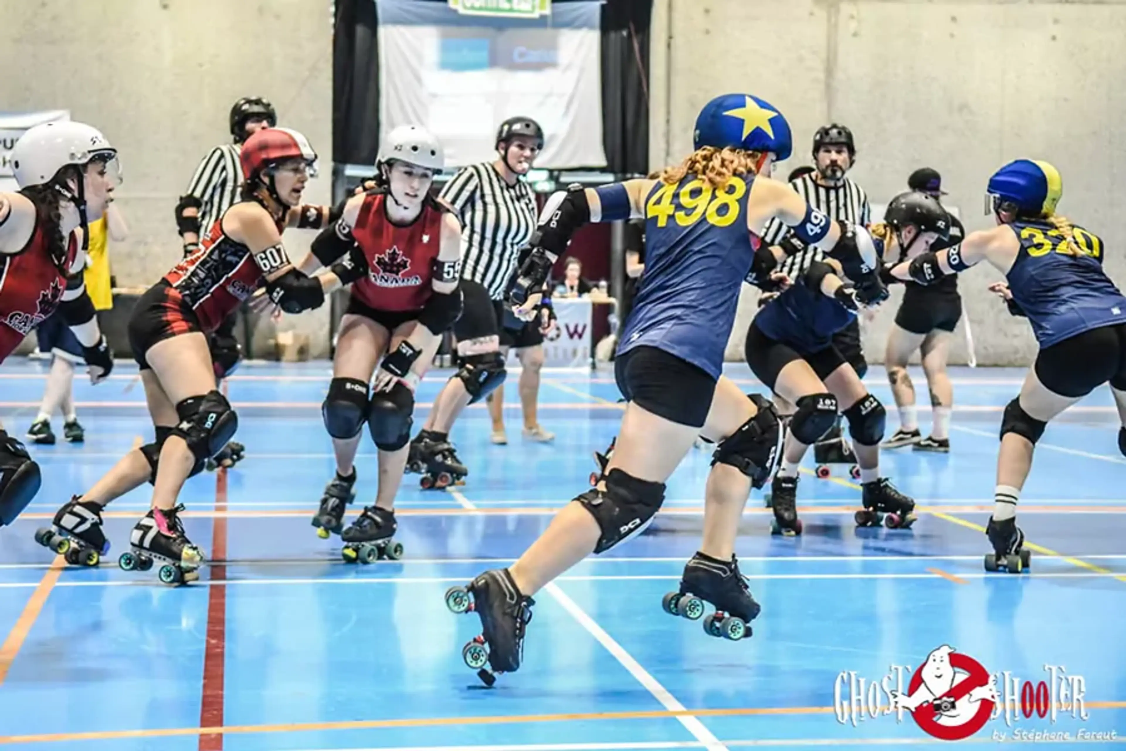 Två lag i rolerderby spelar match inomhus på en blå sportgolvyta, iklädda hjälmar och skyddsutrustning. Domare i randiga tröjor står i bakgrunden och följer spelet noga.