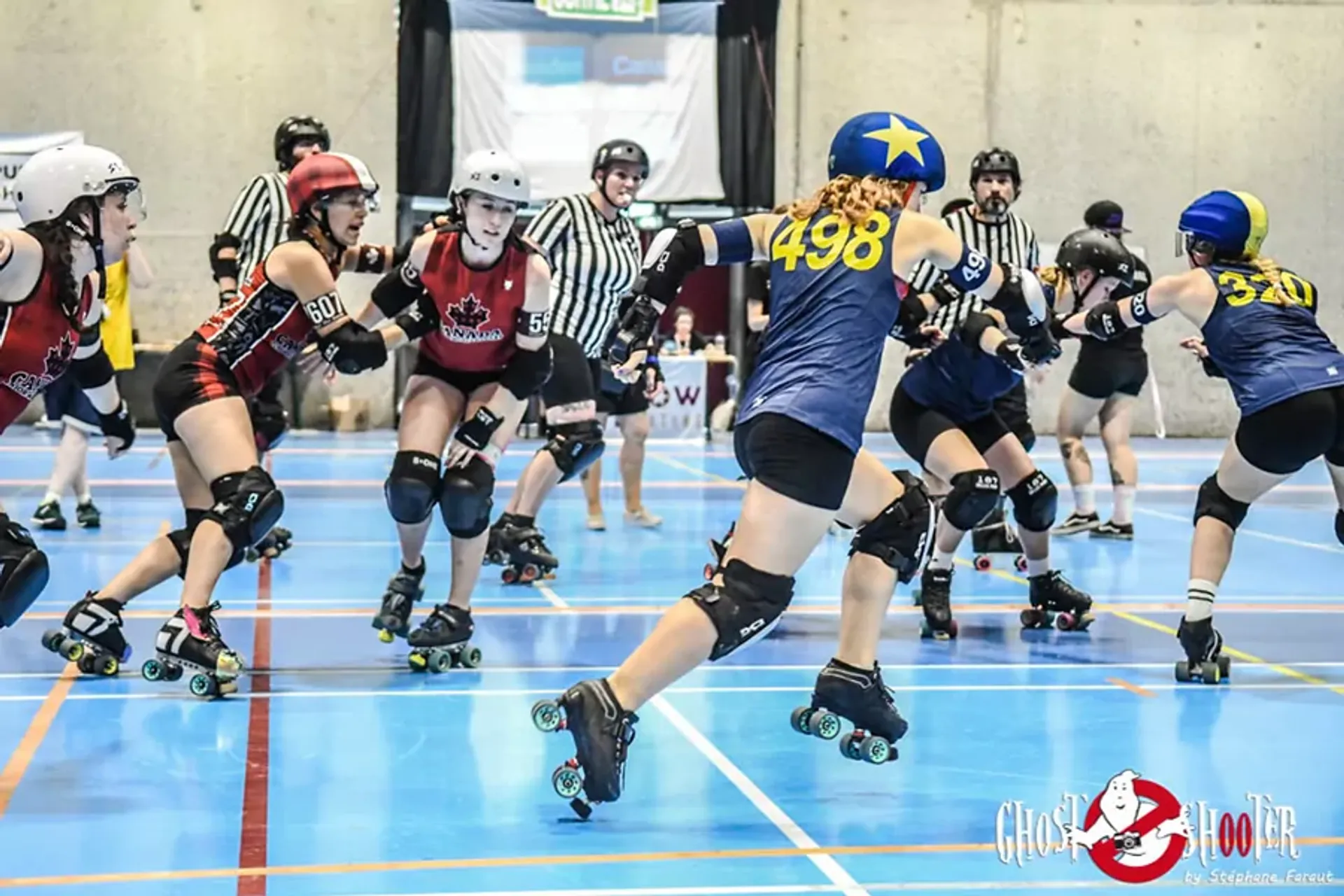Två lag i rolerderby spelar match inomhus på en blå sportgolvyta, iklädda hjälmar och skyddsutrustning. Domare i randiga tröjor står i bakgrunden och följer spelet noga.