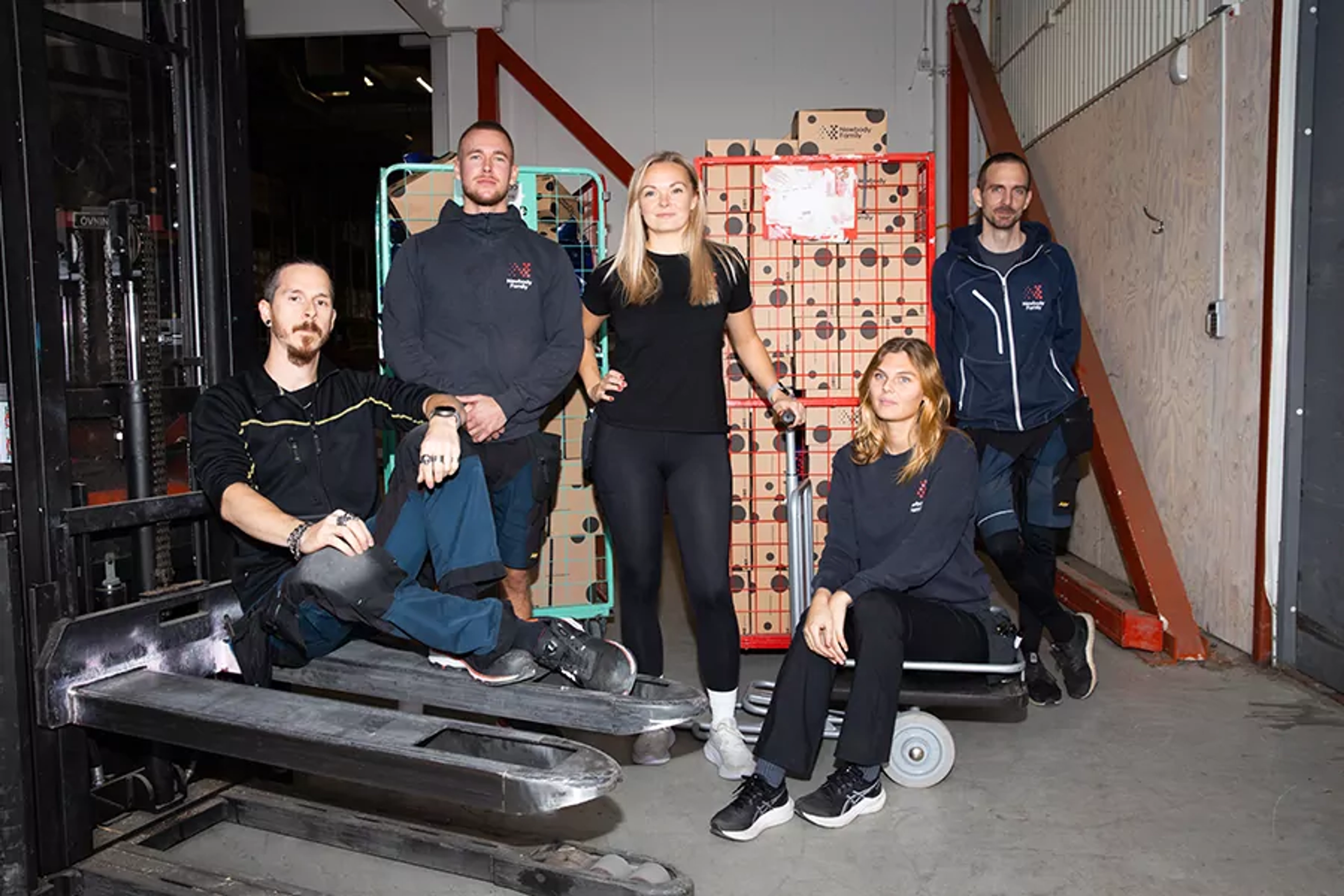 Fem lageranställda samlade runt lagermaterial och transportvagnar i en större industrilokal. Alla bär arbetskläder och vilar en stund under arbetet.