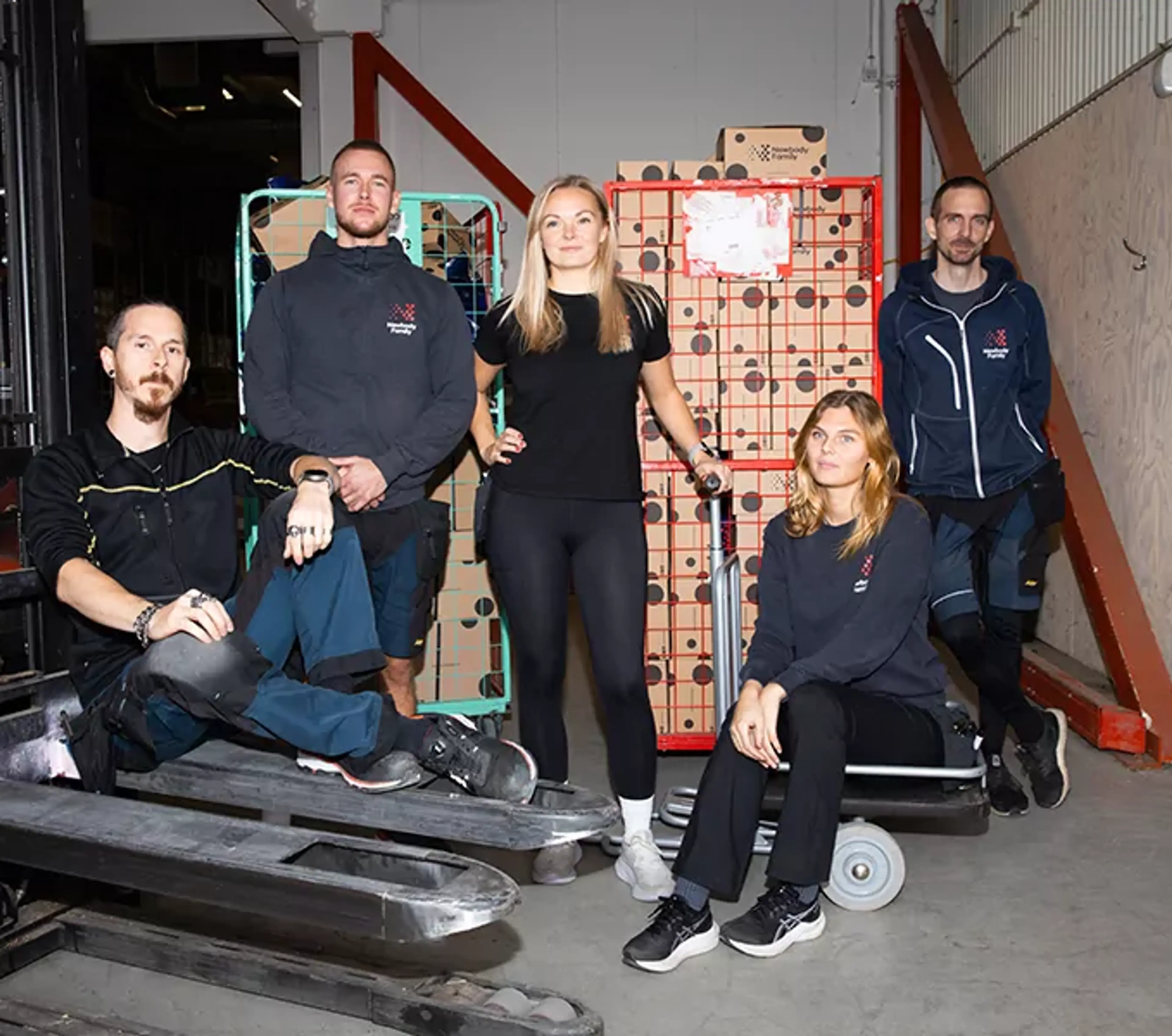 Fem lageranställda samlade runt lagermaterial och transportvagnar i en större industrilokal. Alla bär arbetskläder och vilar en stund under arbetet.