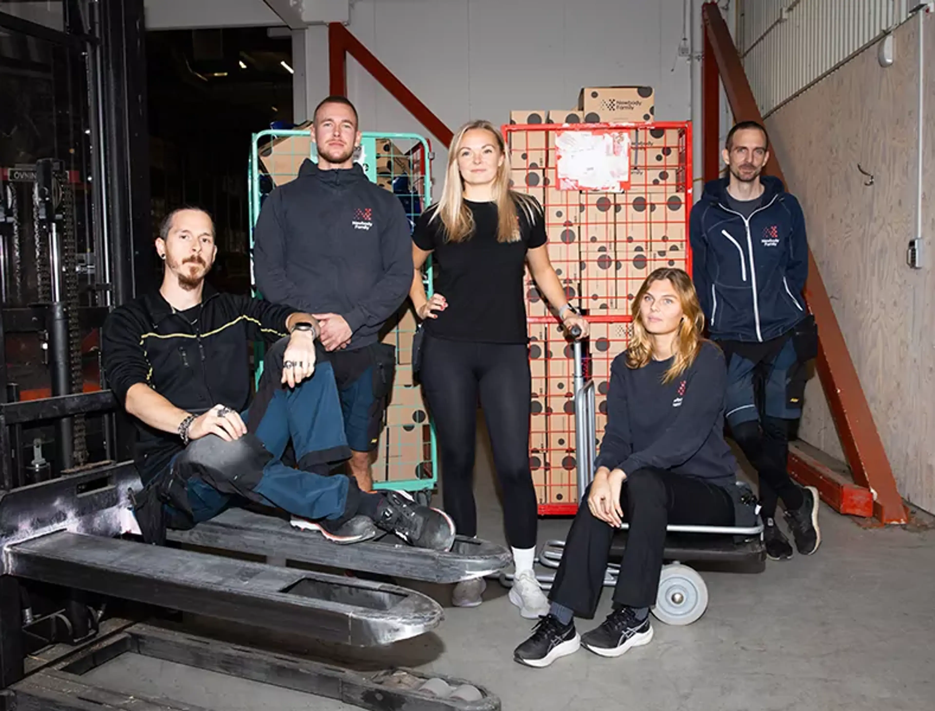 Fem lageranställda samlade runt lagermaterial och transportvagnar i en större industrilokal. Alla bär arbetskläder och vilar en stund under arbetet.