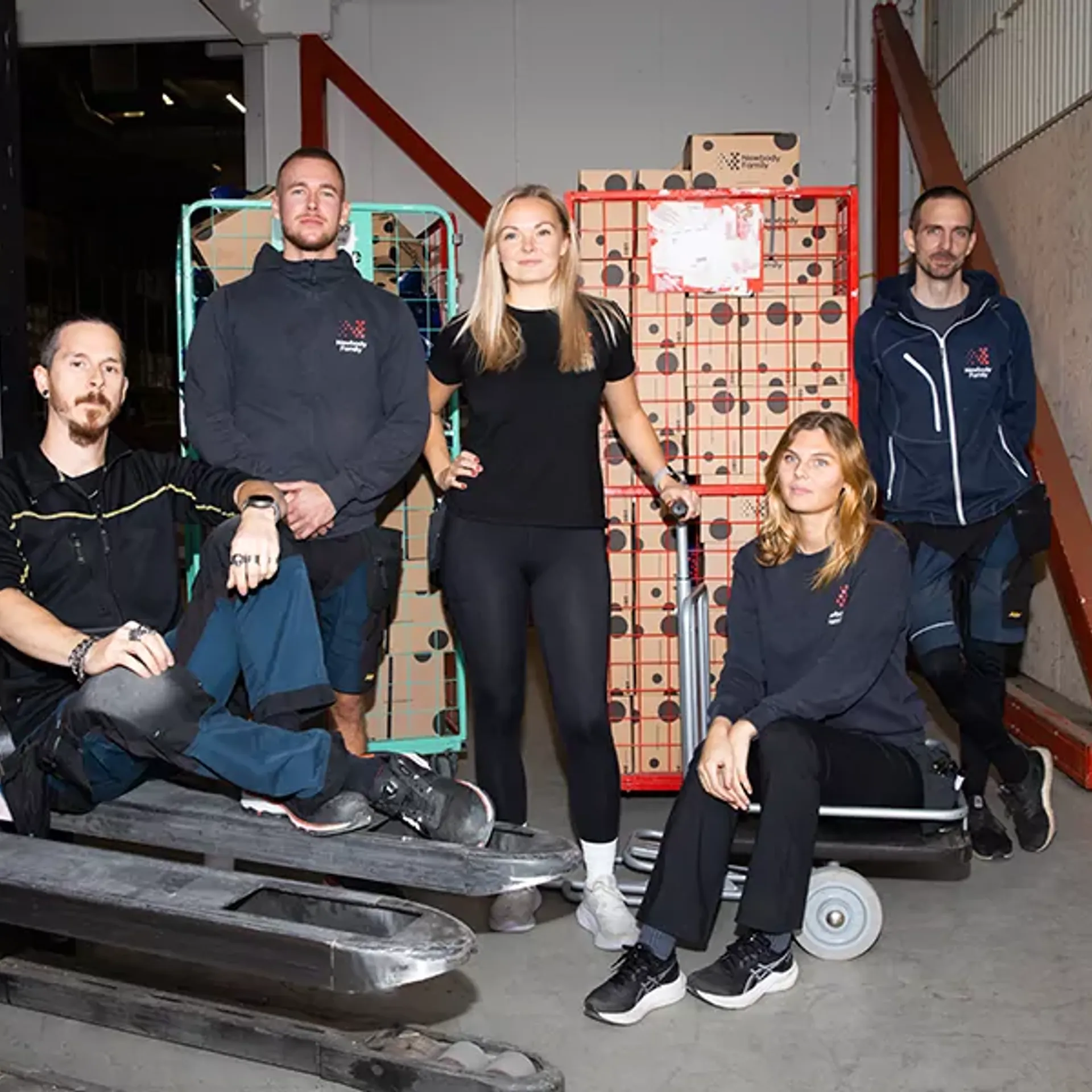 Fem lageranställda samlade runt lagermaterial och transportvagnar i en större industrilokal. Alla bär arbetskläder och vilar en stund under arbetet.