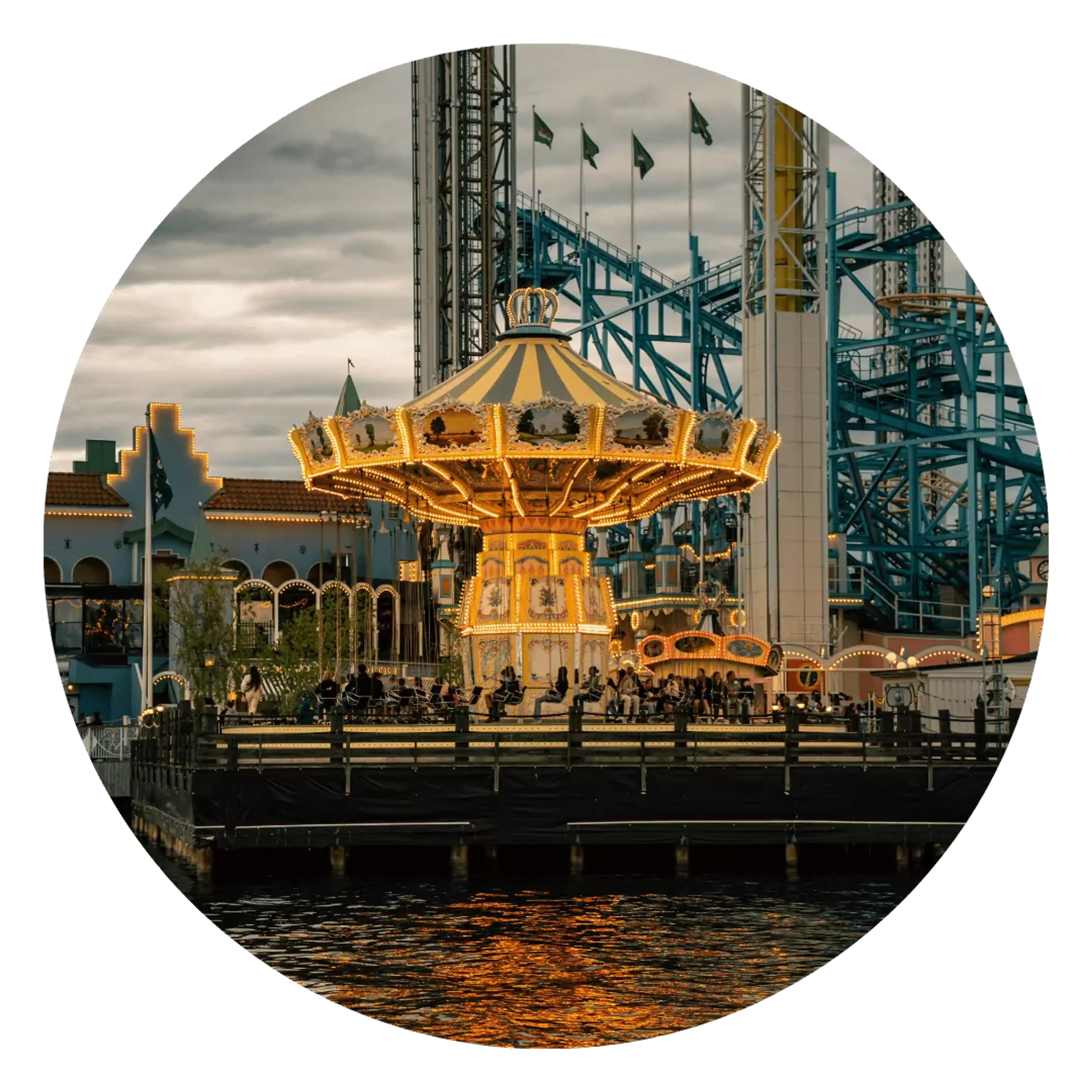 En upplyst karusell med vingsitsar står vid vattnet i en nöjespark under skymning. I bakgrunden syns berg- och dalbanespår i blått och vitt.