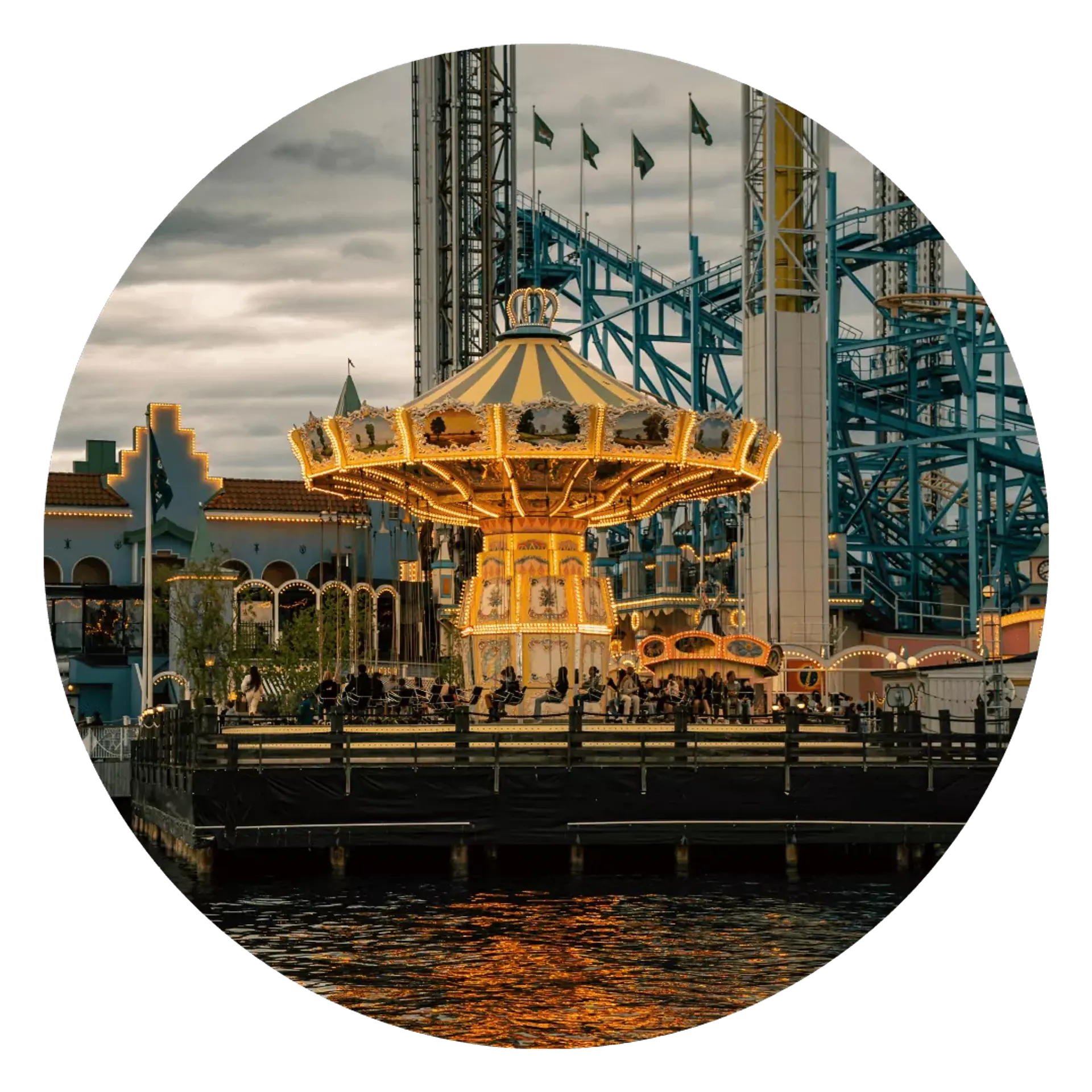 En upplyst karusell med vingsitsar står vid vattnet i en nöjespark under skymning. I bakgrunden syns berg- och dalbanespår i blått och vitt.