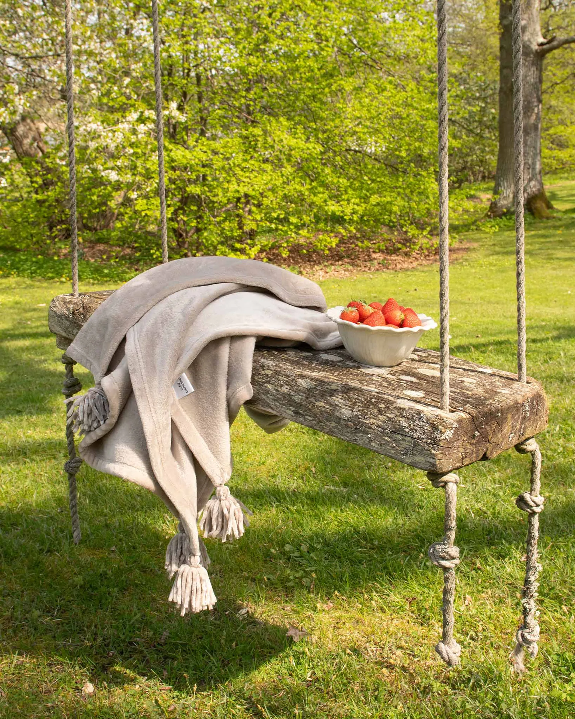 En mjuk, beige fleecefilt med tofsar ligger över en rustik trädyna i en gunga ute i en grönskande trädgård. Bredvid filten står en skål fylld med färska jordgubbar.