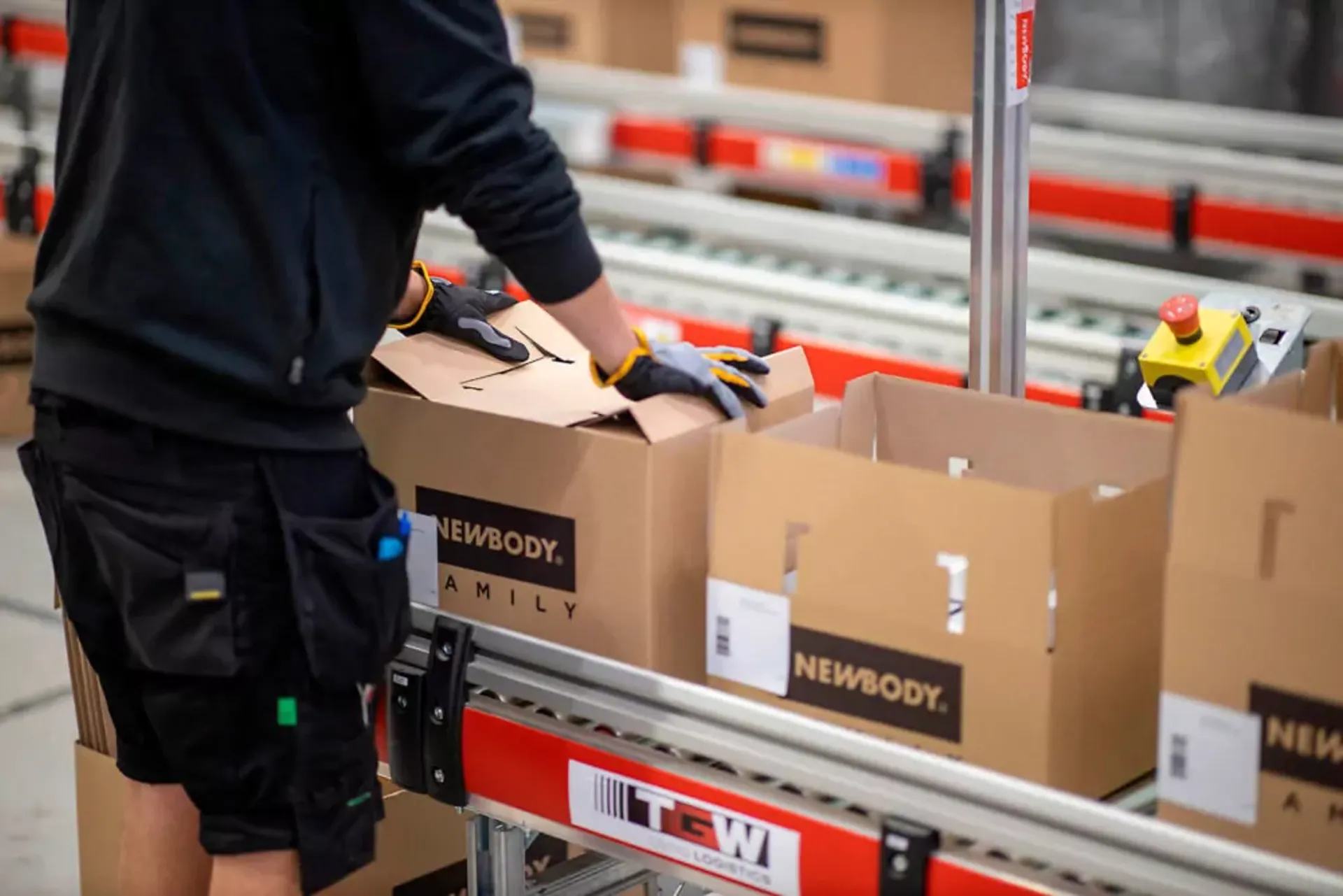 En person med handskar packar kartonger på ett löpande band i en lager- eller packningsmiljö. Kartongerna är märkta med svart text på brun kartong.