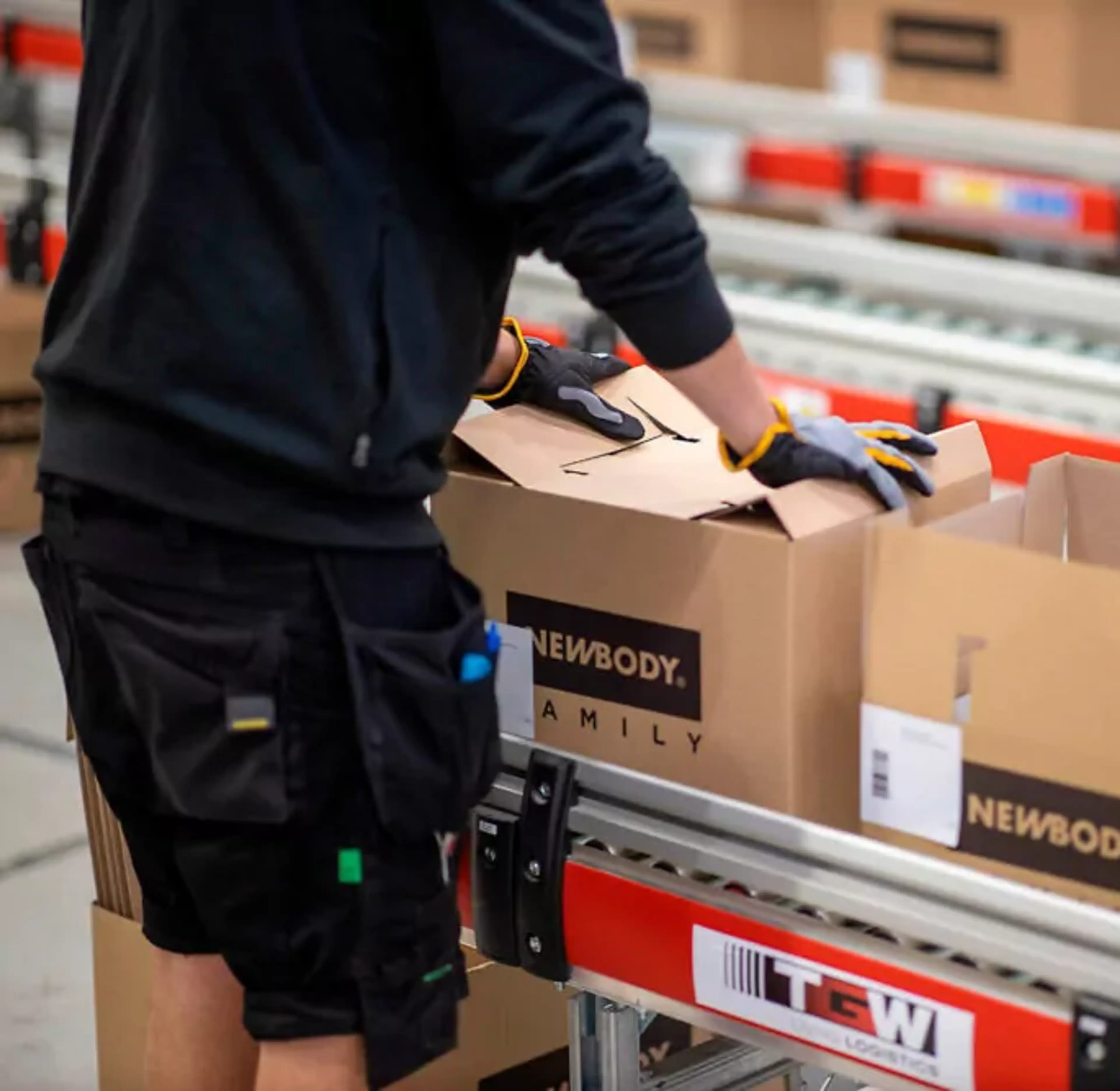 En person med handskar packar kartonger på ett löpande band i en lager- eller packningsmiljö. Kartongerna är märkta med svart text på brun kartong.