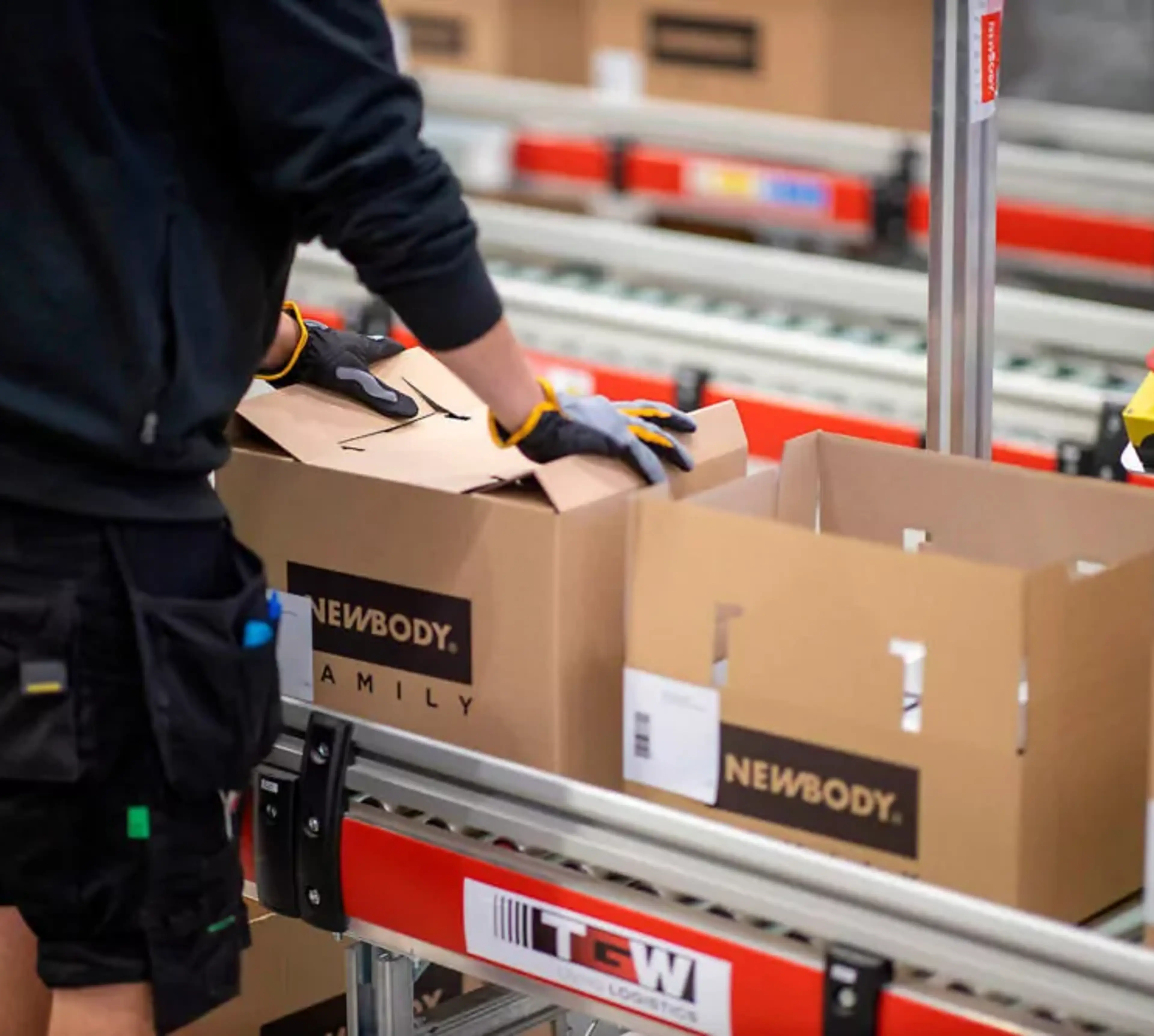 En person med handskar packar kartonger på ett löpande band i en lager- eller packningsmiljö. Kartongerna är märkta med svart text på brun kartong.