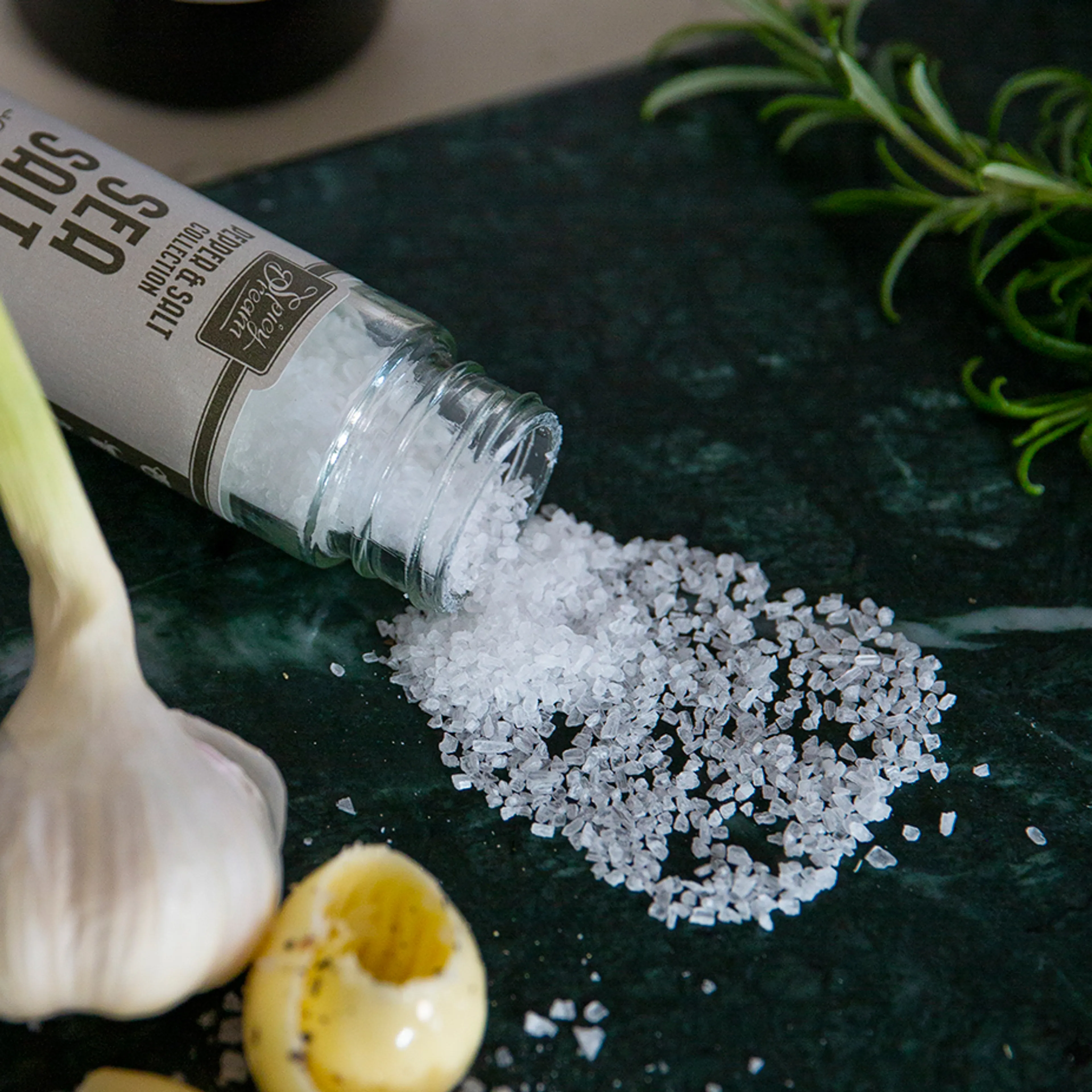 Ett glasrör med grovt havssalt som är urtappat på en mörk grön marmorbänk. I bakgrunden syns en vitlök, vitlöksskivor och en kvist rosmarin.
