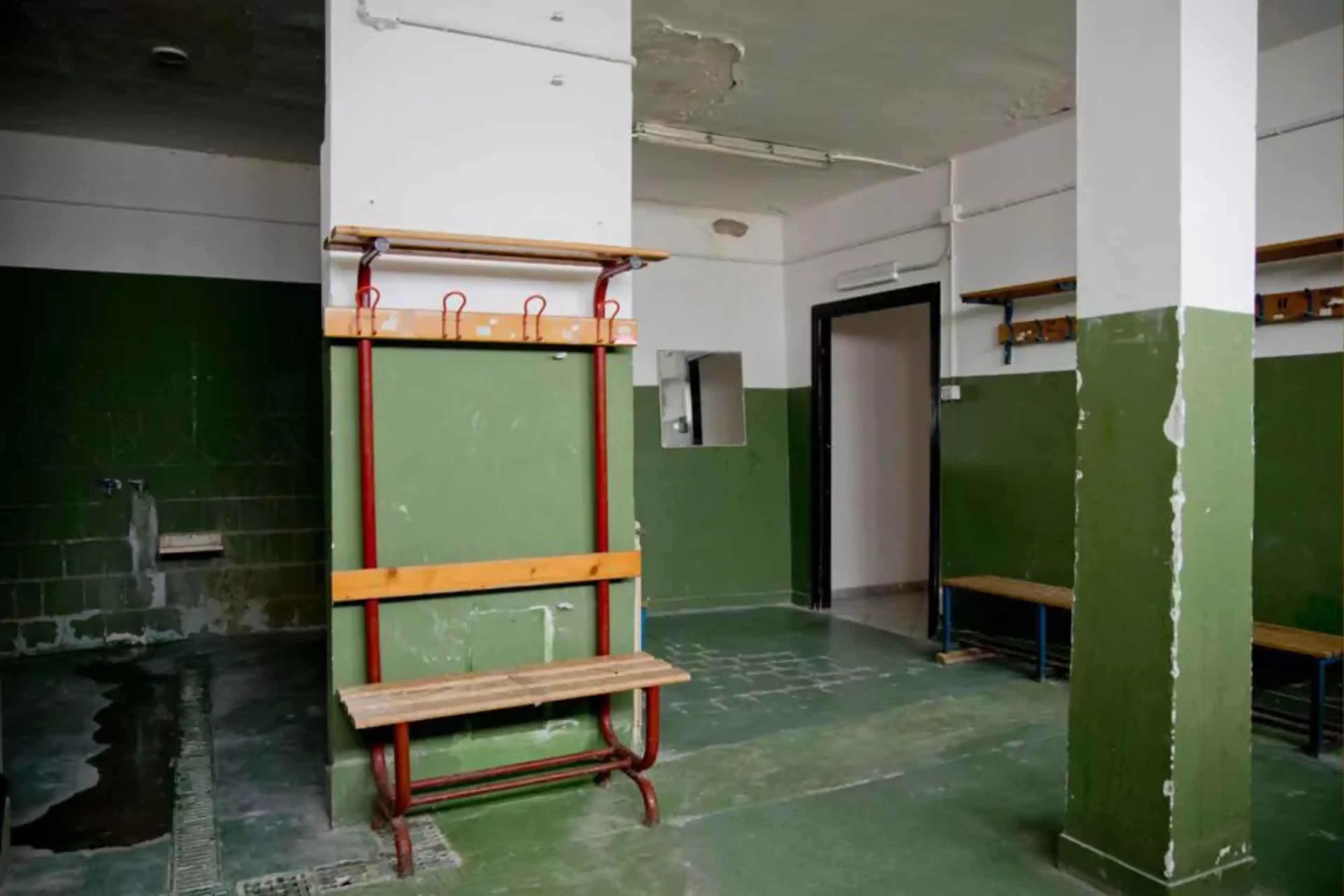 Ett omklädningsrum med gröna väggar, träsittbänkar och enkla klädhängare. Rummet känns rustikt med slitna ytor och en funktionell, sparsmakad inredning.