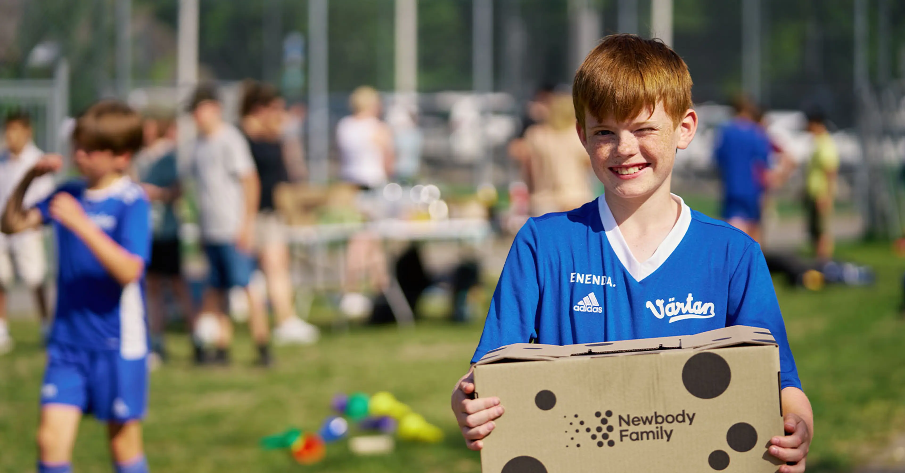 En pojke i blå fotbollströja håller en kartong med Newbody Family-logotypen på en grön gräsmatta. I bakgrunden syns flera barn och ett bord med olika saker.