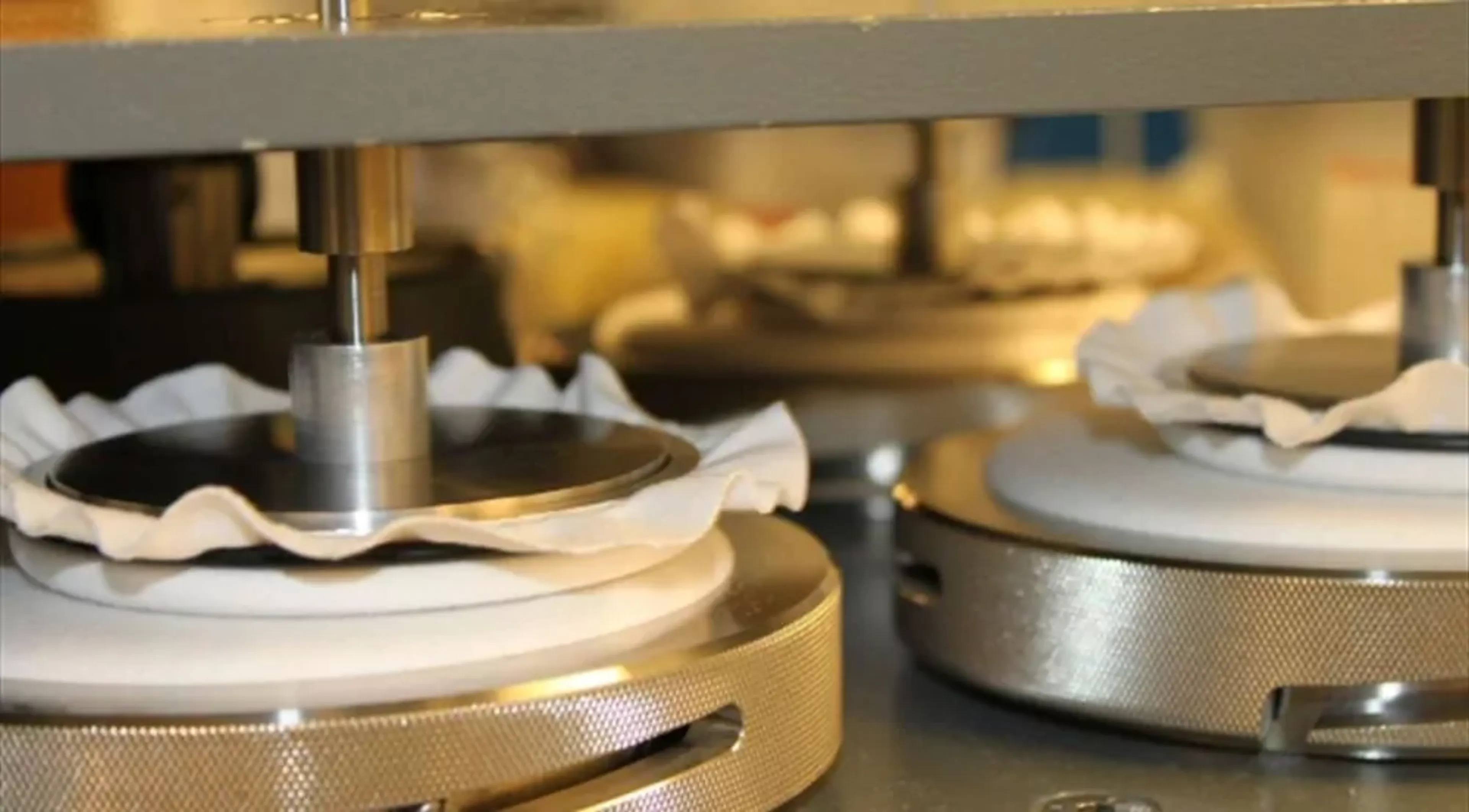Närbild på en maskin som pressar tygstycken mellan metallplattor, troligen för provningar eller test av material. Textilen är vit och nestad i maskinens runda delar.