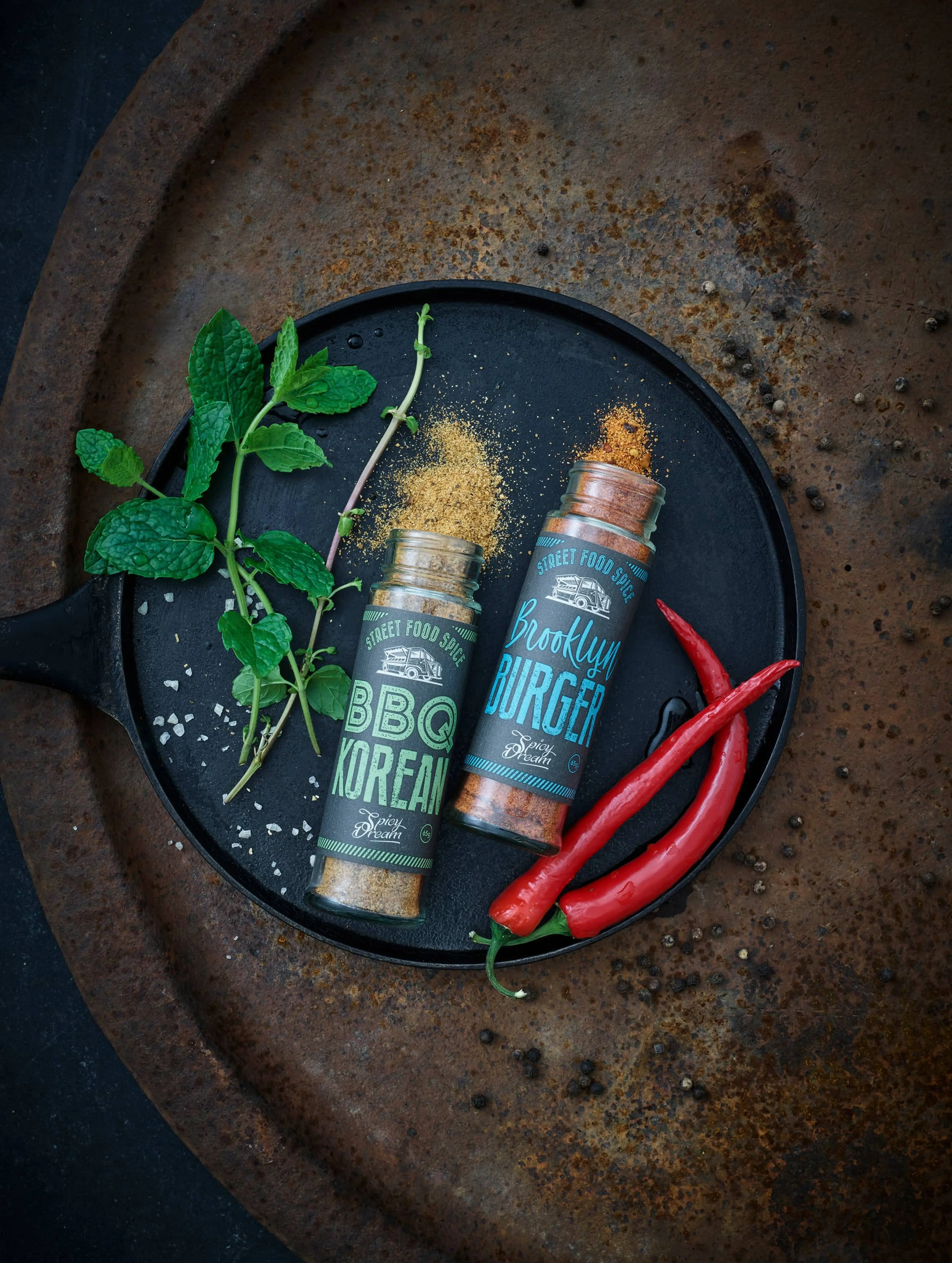 Två kryddburkar med smakrik bbq och burger-krydda tillsammans med färska örter och röda chilipeppar på en gjutjärnspanna. Smulade kryddor ligger utspridda runt burkarna.