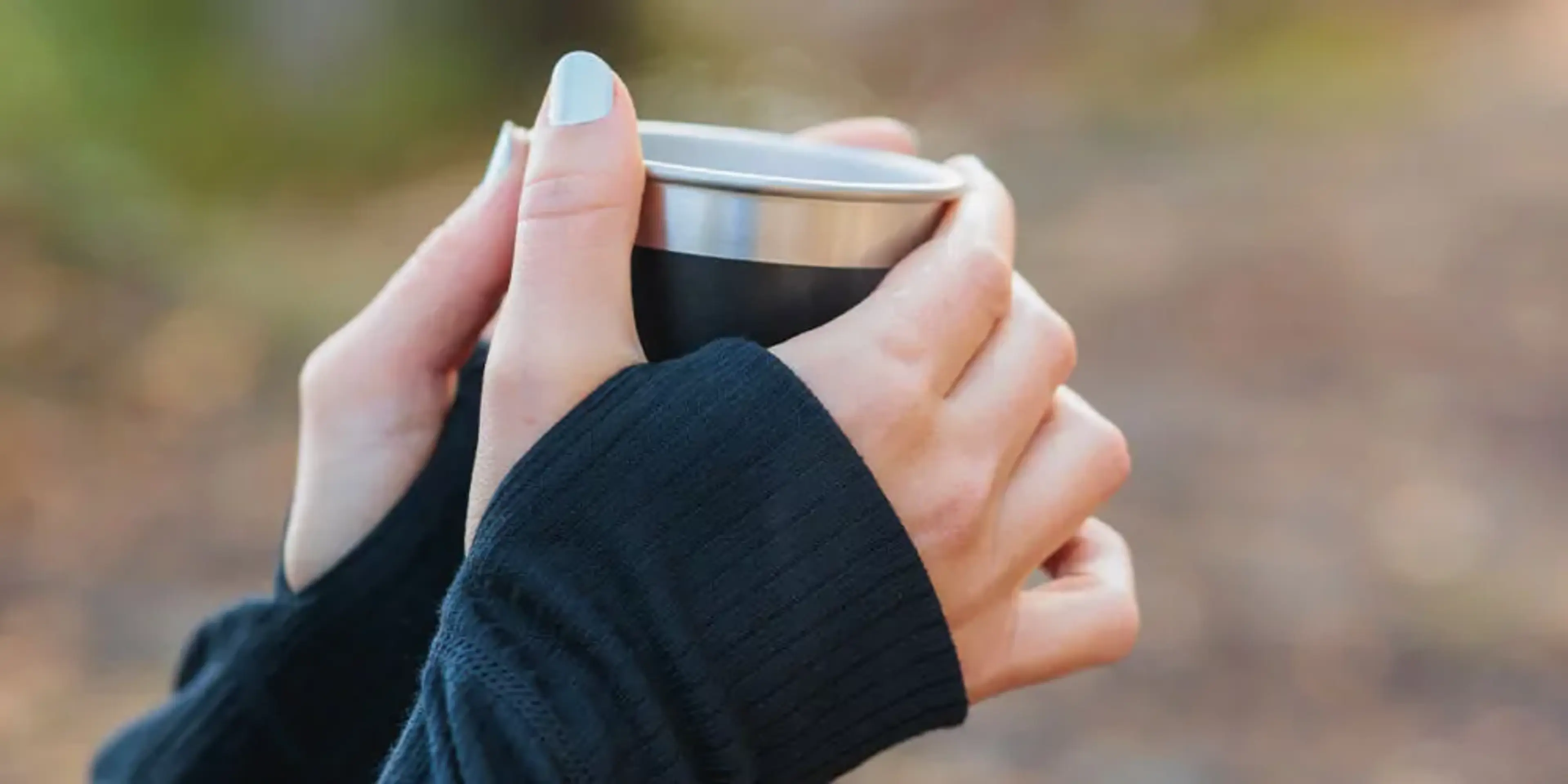 Två händer i svarta ärmar håller en metallmugg med lock, utomhus i mjukt ljus. Muggen ser ut att vara isolerad för varma drycker.