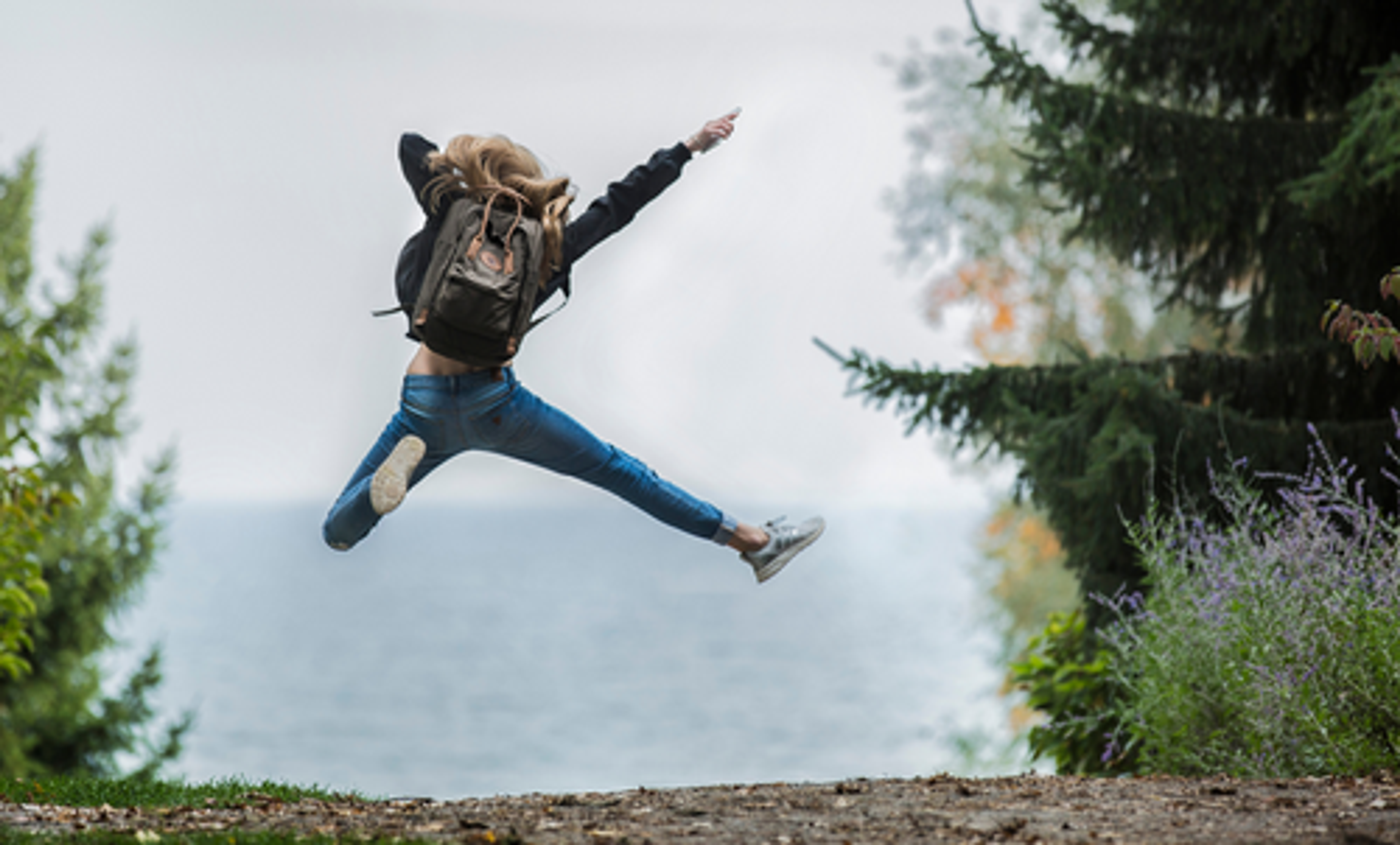 En person i jeans och svart jacka hoppar glatt med utsträckta armar och ben på en skogsstig med havet i bakgrunden. Runtomkring syns gröna träd och buskar.