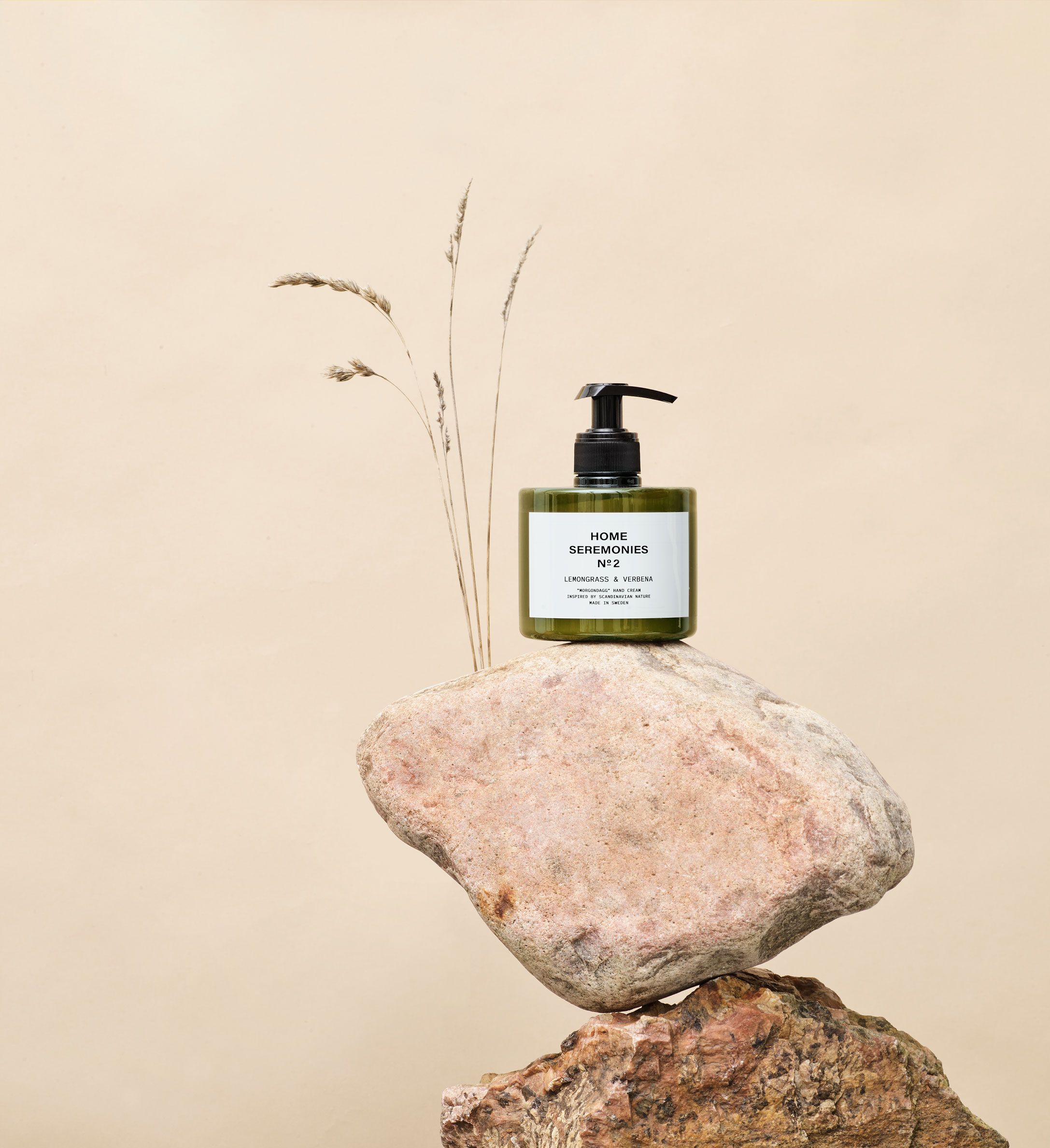 En grön plastflaska med pump innehållande handkräm står balanserad på två stenblock, med några torkade grästjärnor i bakgrunden.