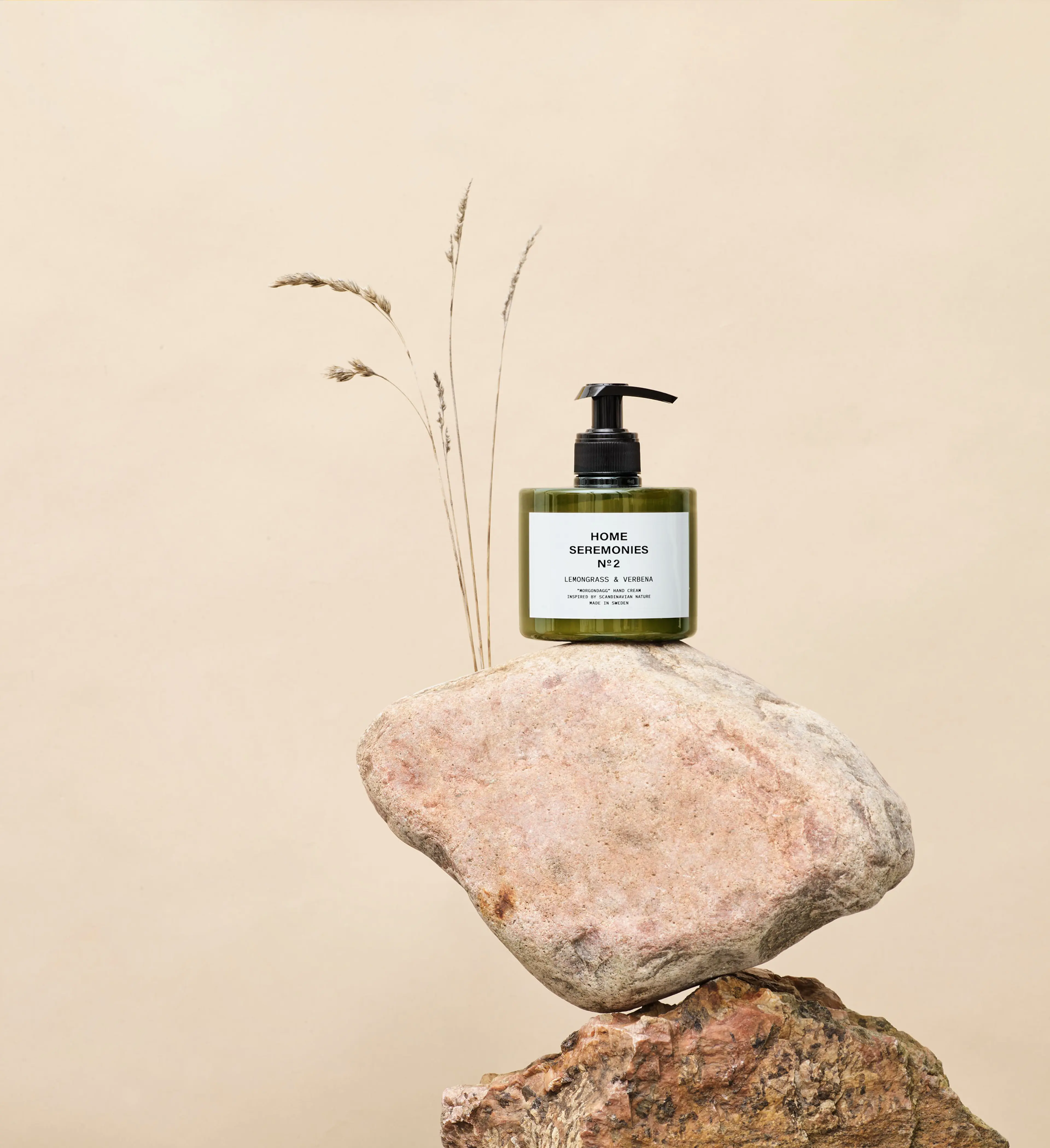 En grön plastflaska med pump innehållande handkräm står balanserad på två stenblock, med några torkade grästjärnor i bakgrunden.