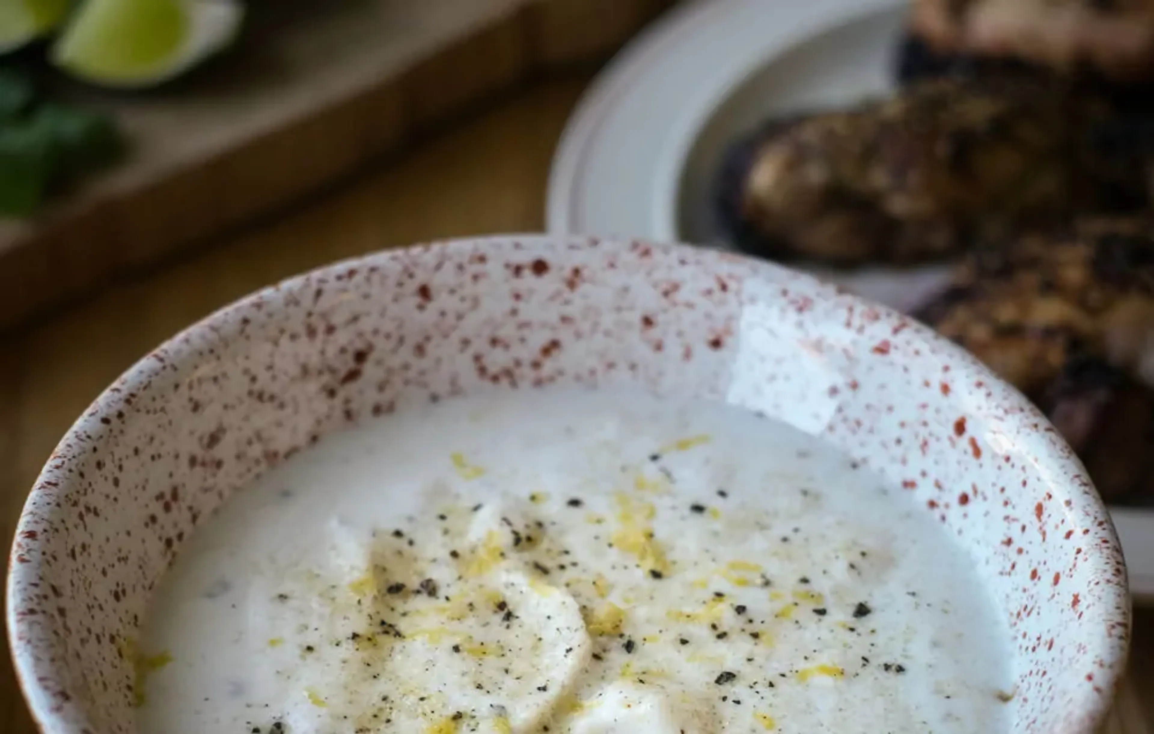 En skål med krämig citronsås, toppad med nymalen svartpeppar och citronzest. I bakgrunden skymtar en tallrik med grillat kött.
