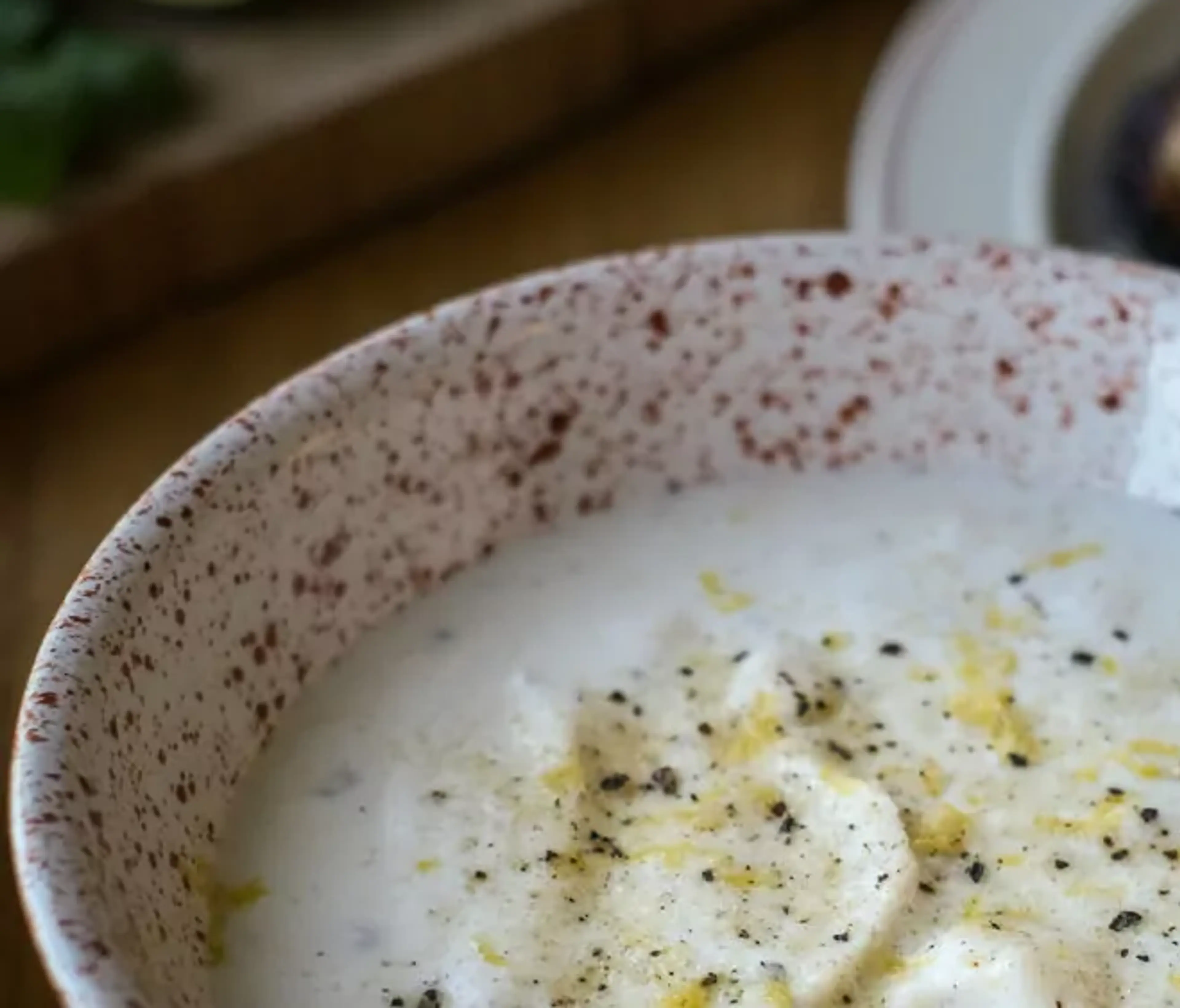 En skål med krämig citronsås, toppad med nymalen svartpeppar och citronzest. I bakgrunden skymtar en tallrik med grillat kött.