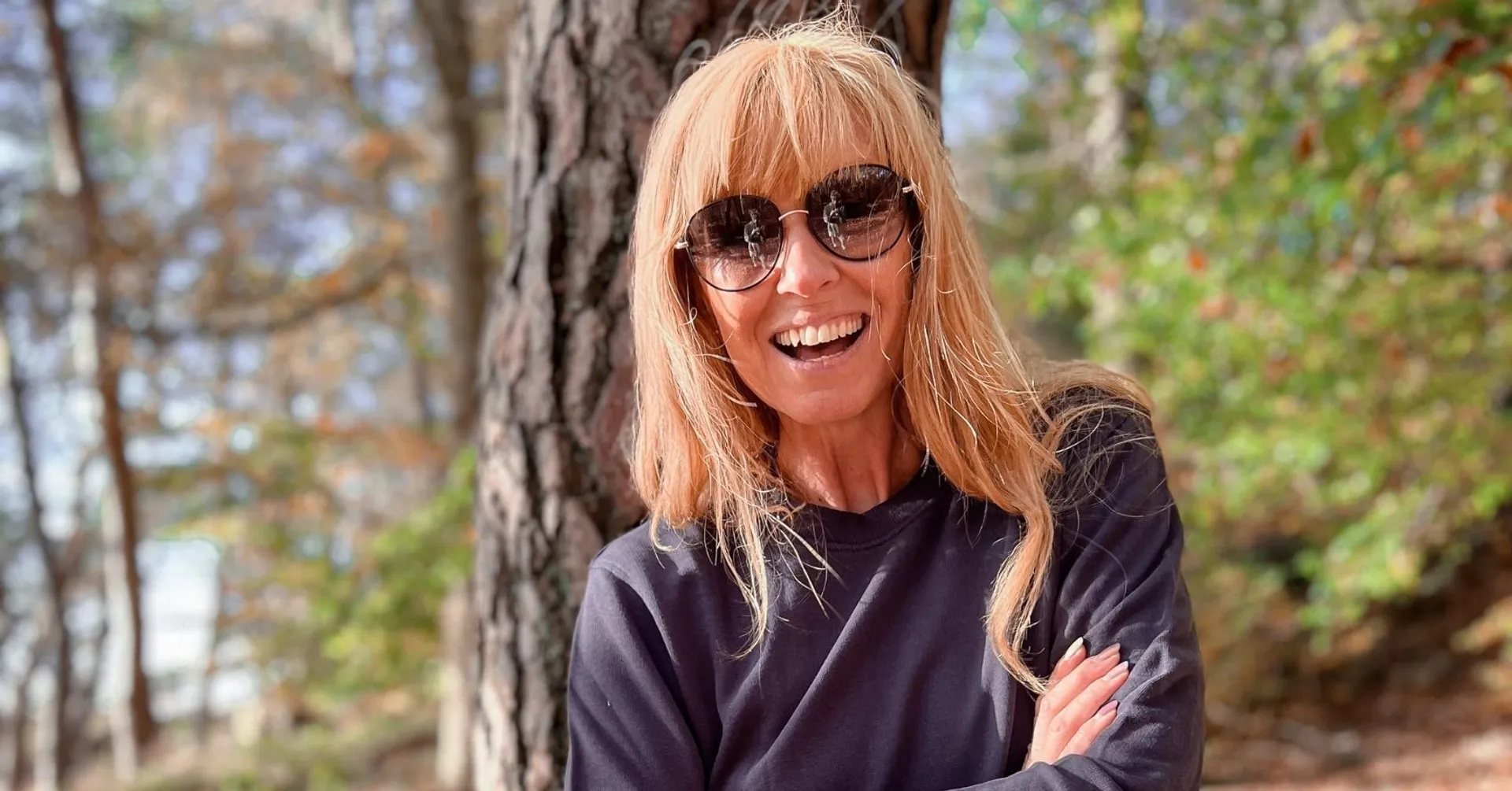 En kvinna med solglasögon och långt blont hår står med armarna i kors framför en trädstam i en skogsmiljö. Hon bär en mörk, långärmad tröja och ler mot kameran.