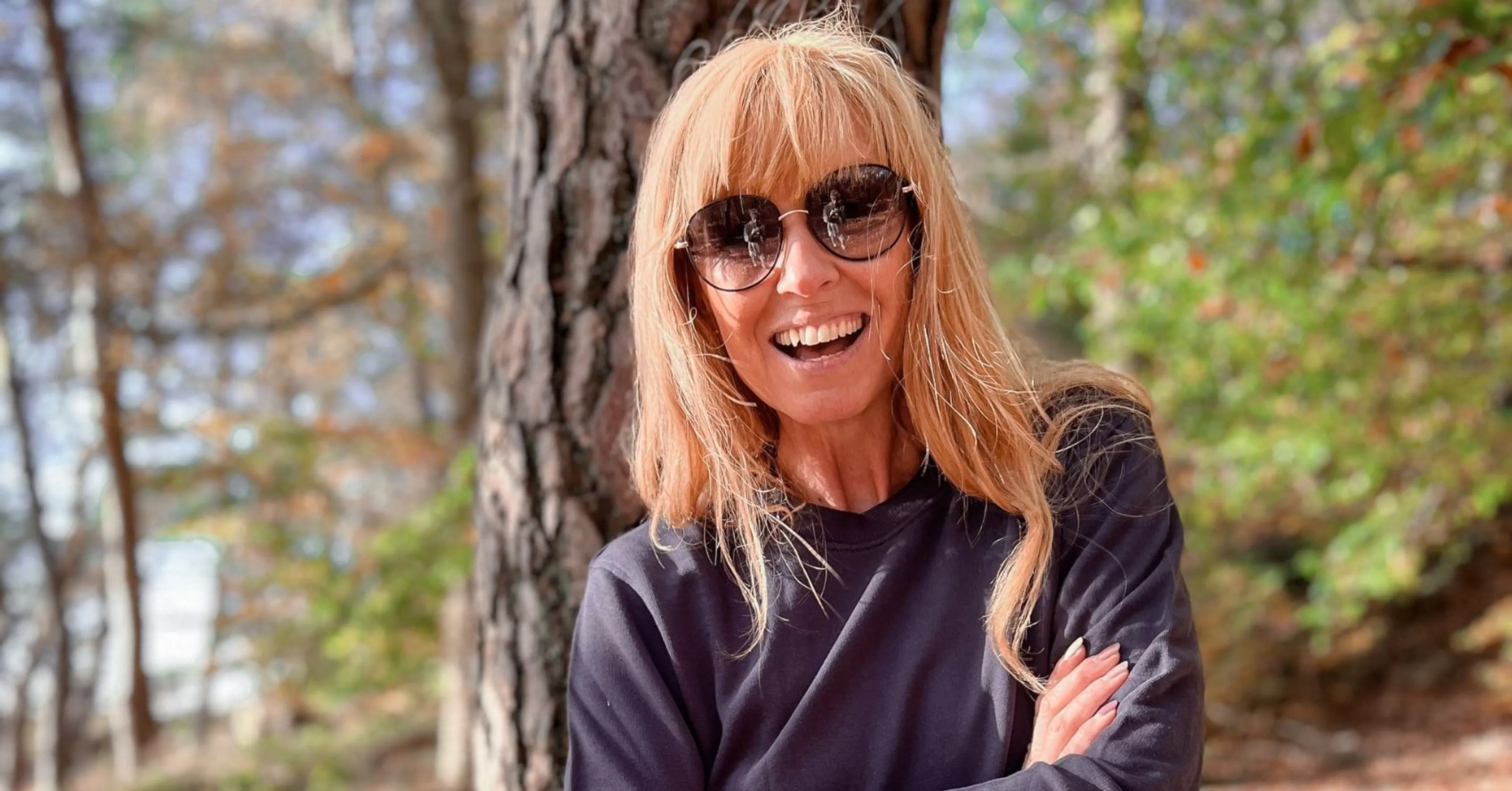 En kvinna med solglasögon och långt blont hår står med armarna i kors framför en trädstam i en skogsmiljö. Hon bär en mörk, långärmad tröja och ler mot kameran.