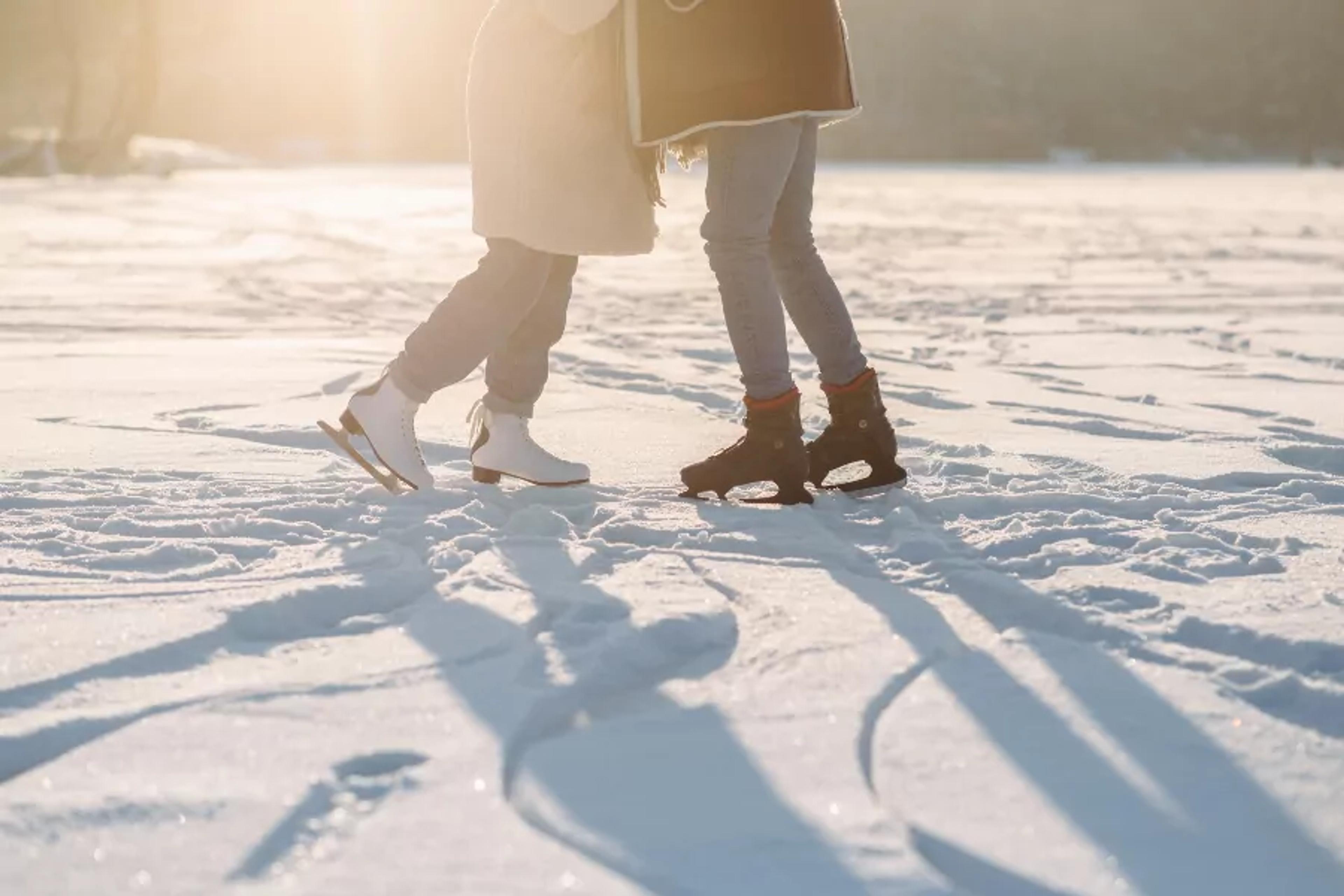 Två personer står på snöklädd is med skridskor på, en med vita och en med svarta skridskor. Solen kastar långa skuggor över isen.
