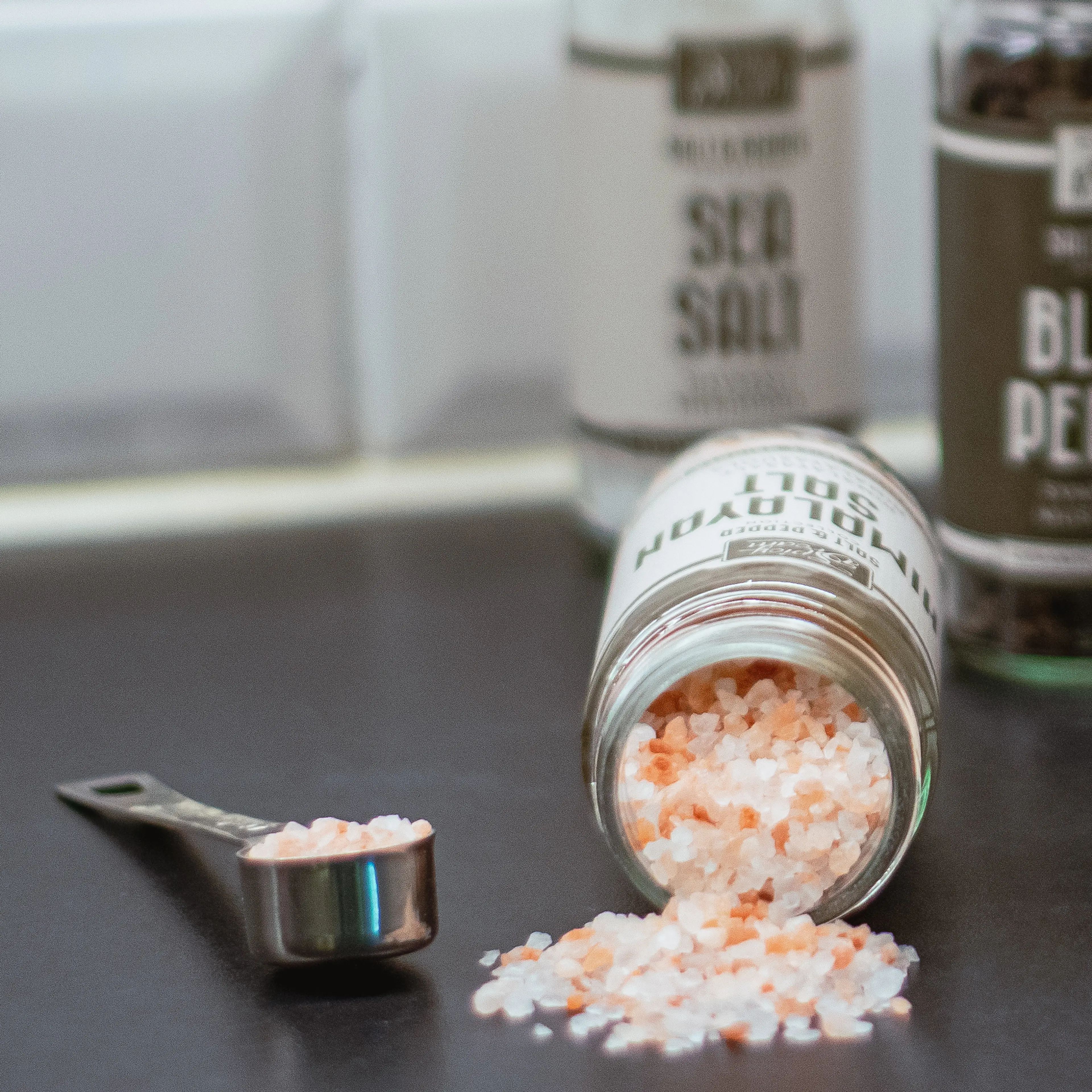 Ett glasburk ligger på sidan med rosa och vita himalayasaltkorn som spillts ut på en mörk yta, bredvid en liten måttsked fylld med salt. I bakgrunden syns burkar med kryddor.
