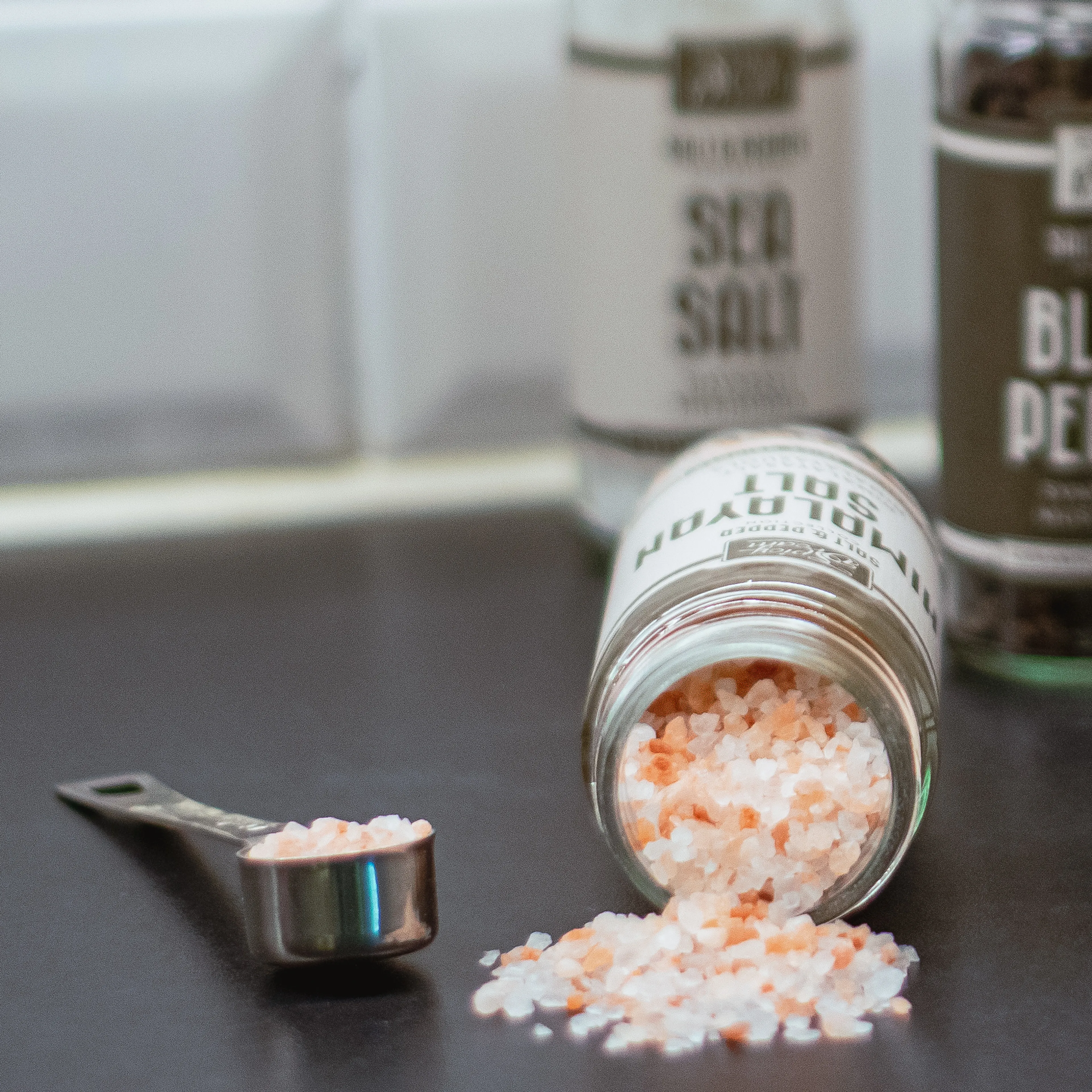 Ett glasburk ligger på sidan med rosa och vita himalayasaltkorn som spillts ut på en mörk yta, bredvid en liten måttsked fylld med salt. I bakgrunden syns burkar med kryddor.