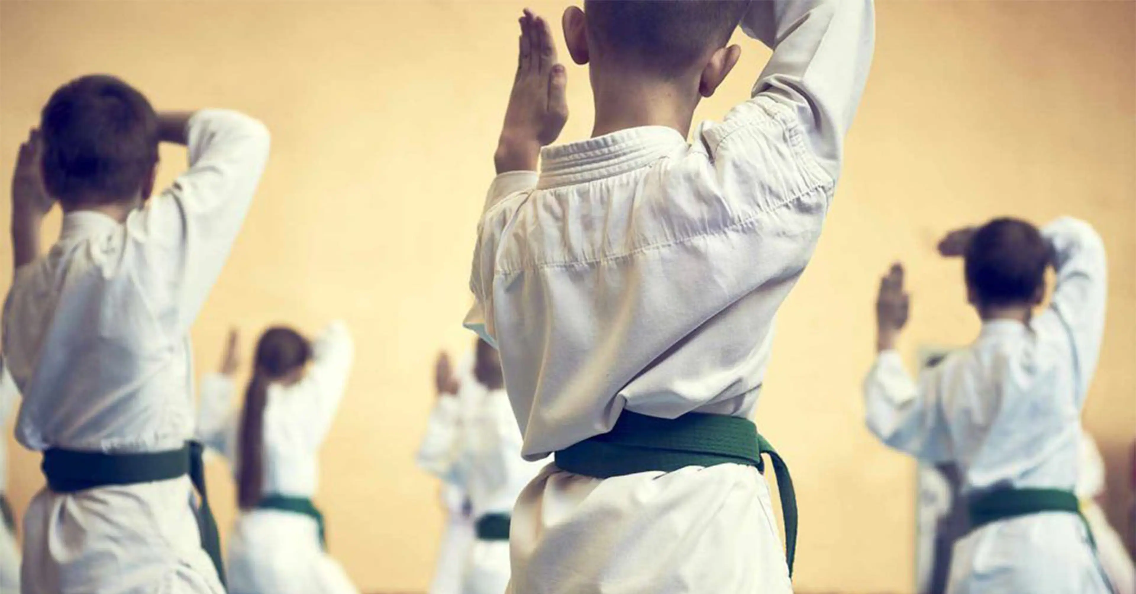 Barn i vita dräkter med gröna bälten tränar kampsport i en dojo. Gruppen fokuserar på en gemensam rörelse med händerna höjda.