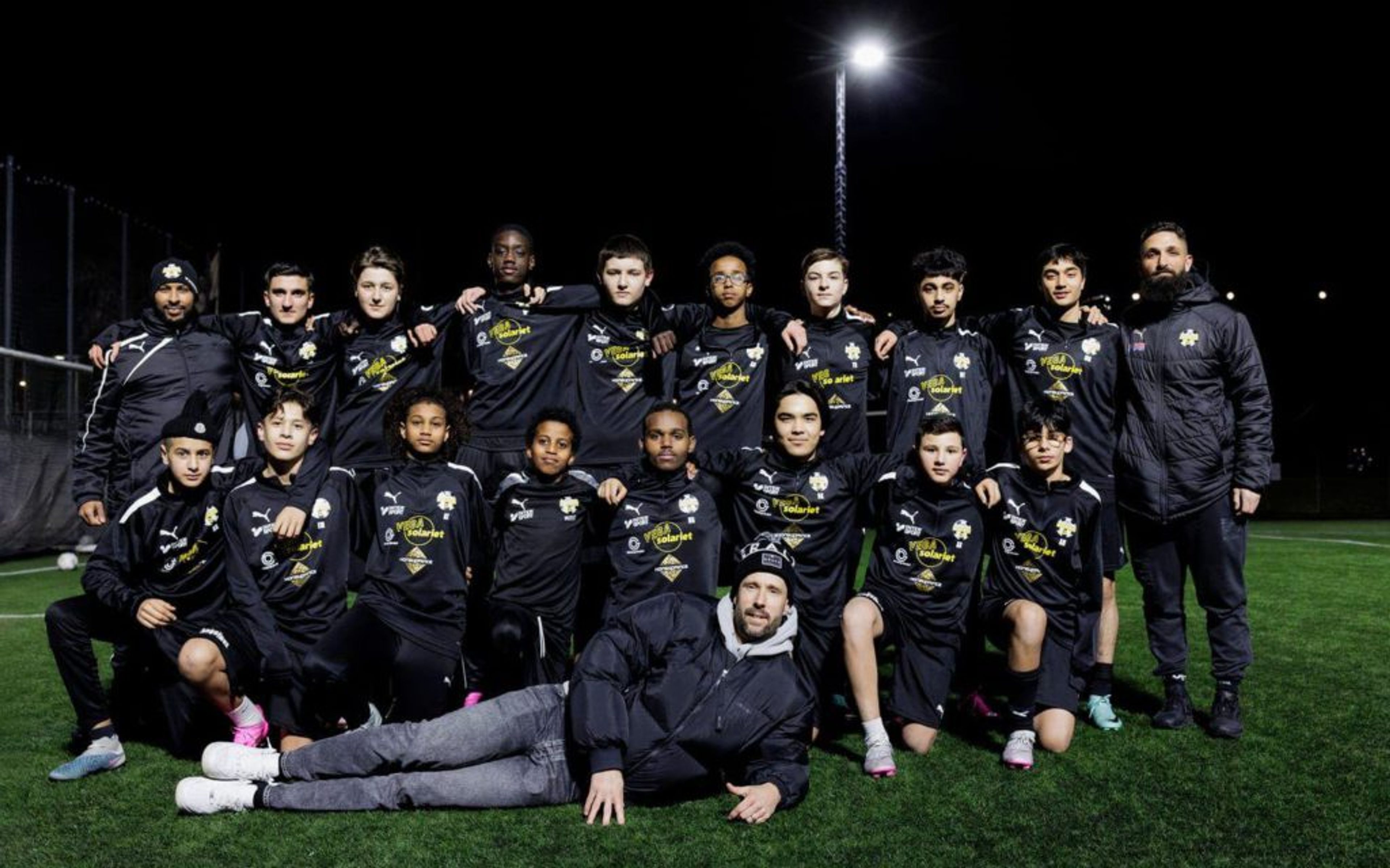 Ungdomslag i fotboll samlat på grön konstgräsplan under kvällstid, klädda i svarta matchställ med logotyper. En person ligger avslappnat framför laget medan resten står och sitter i två rader.