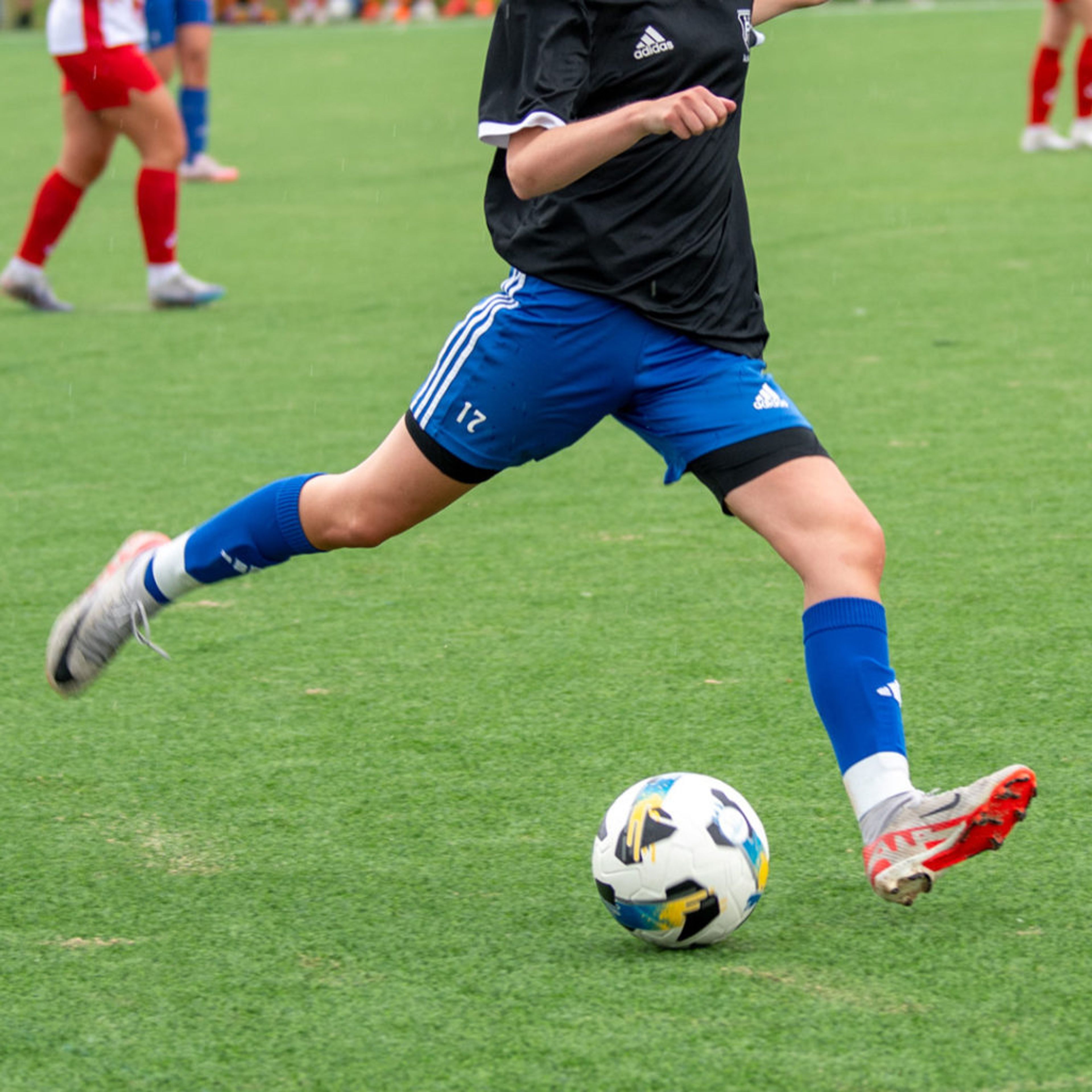 En person i blåa shorts och blåa strumpor sparkar en vit fotboll på en grön gräsplan. Andra spelare syns i bakgrunden.