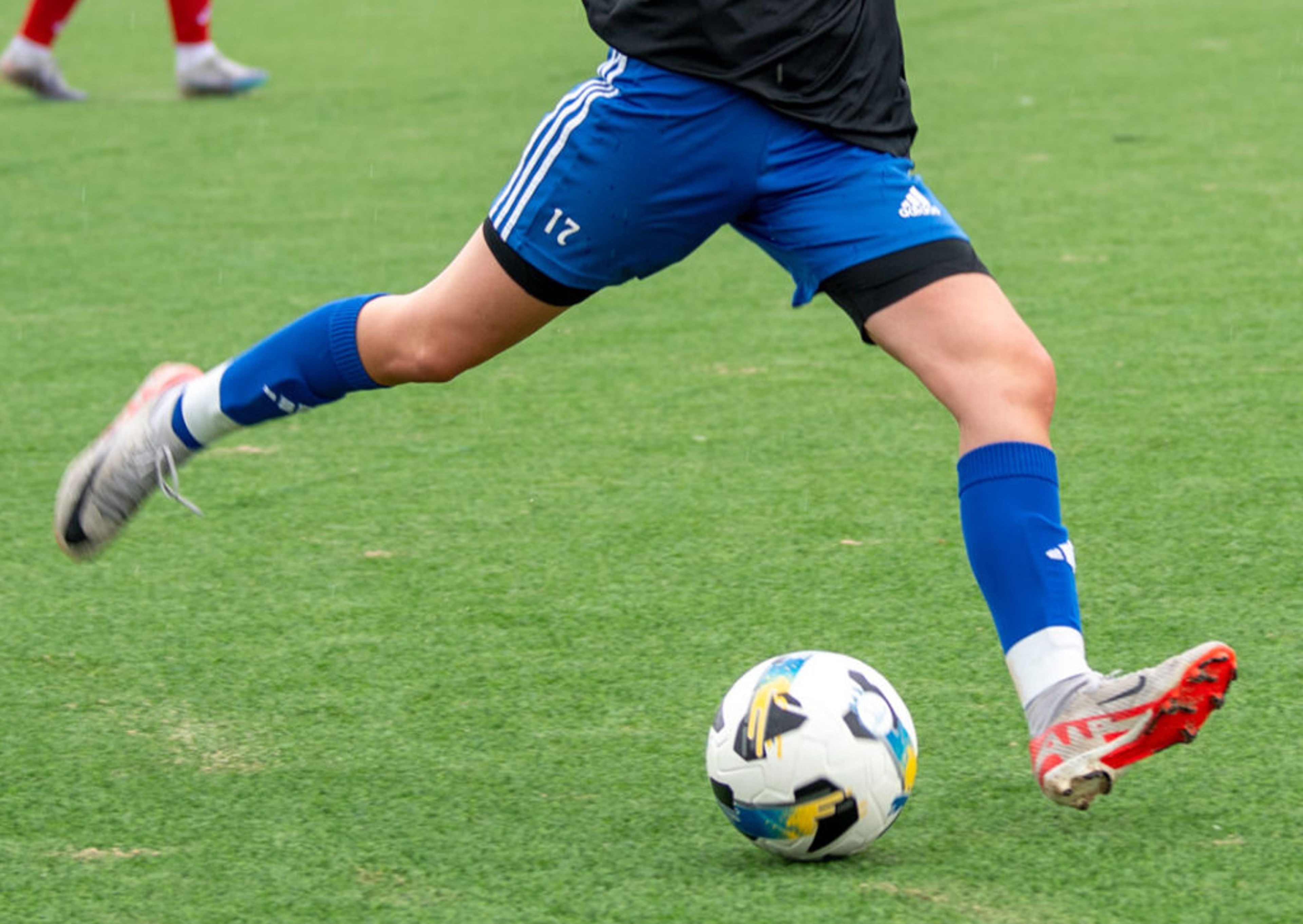En person i blåa shorts och blåa strumpor sparkar en vit fotboll på en grön gräsplan. Andra spelare syns i bakgrunden.