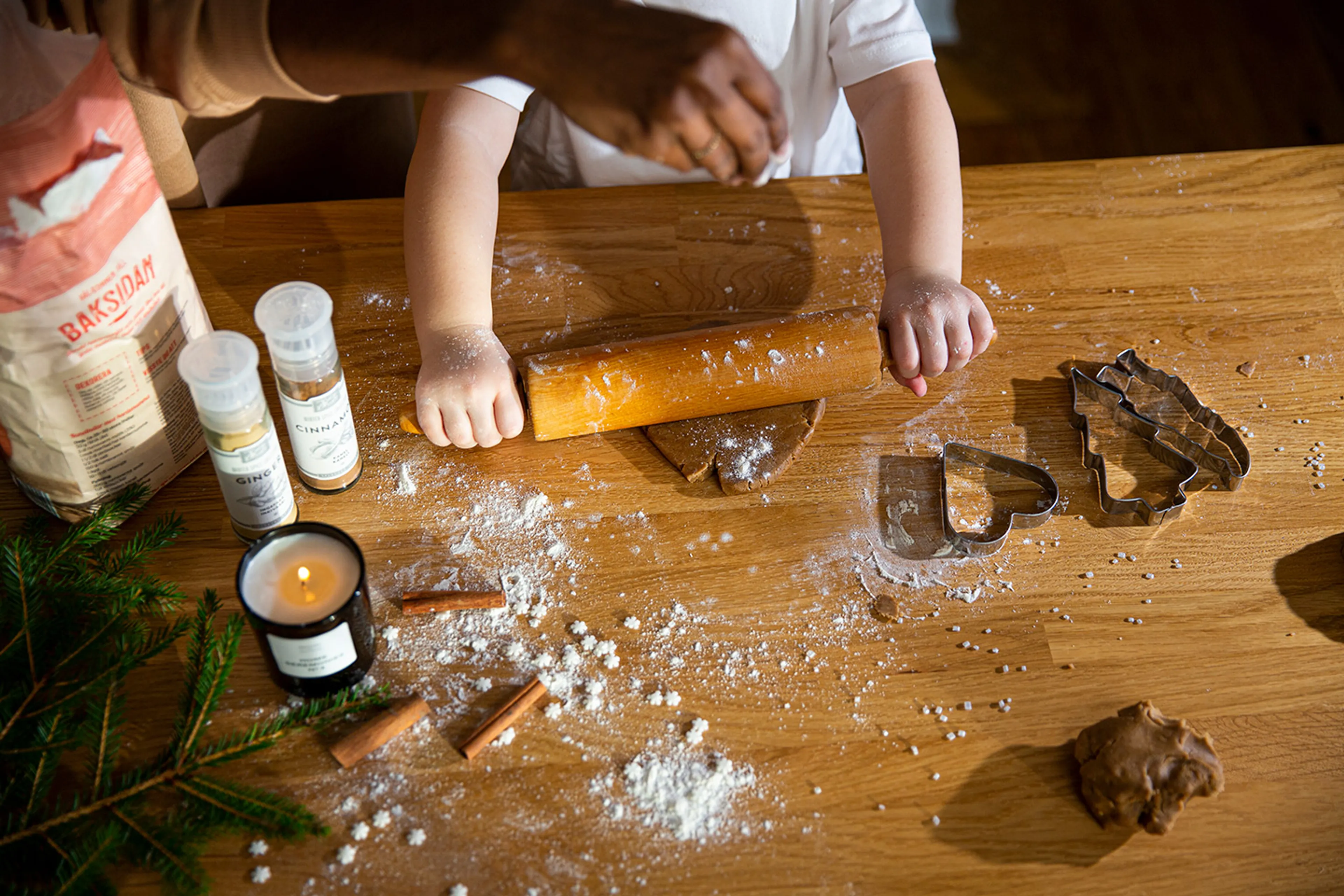 Ett barn kavlar pepparkaksdeg på ett träbord med formar i hjärt- och granform bredvid. Bordet är täckt av mjöl och kryddor som kanel, ingefära och kanel.
