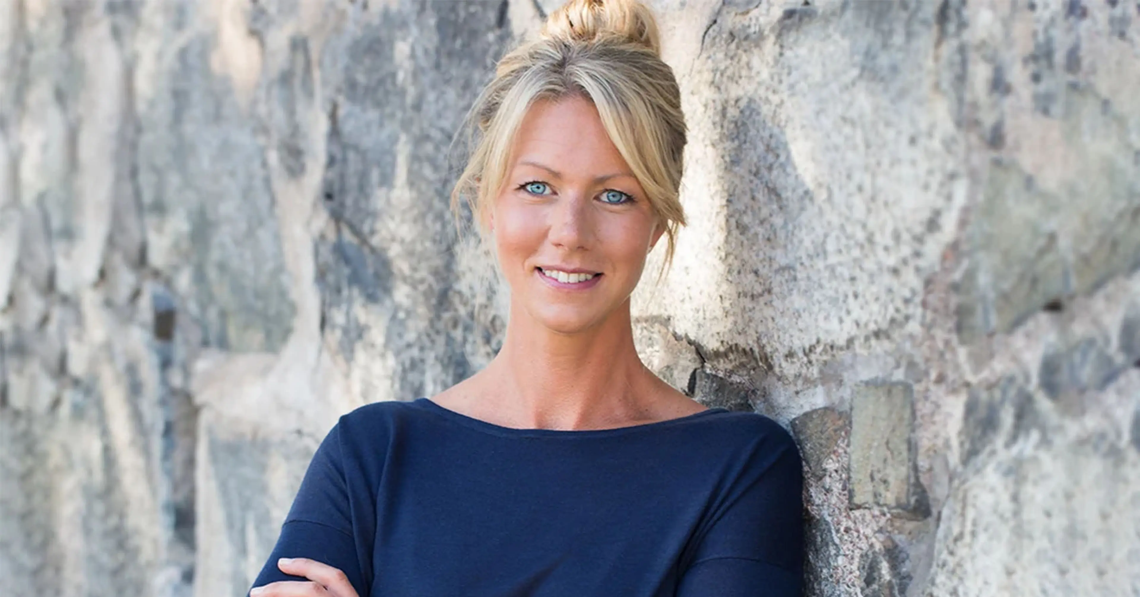 Kvinna med ljusblont hår uppsatt i knut, klädd i en mörkblå tröja, står lutad mot en stenmur med armarna i kors.