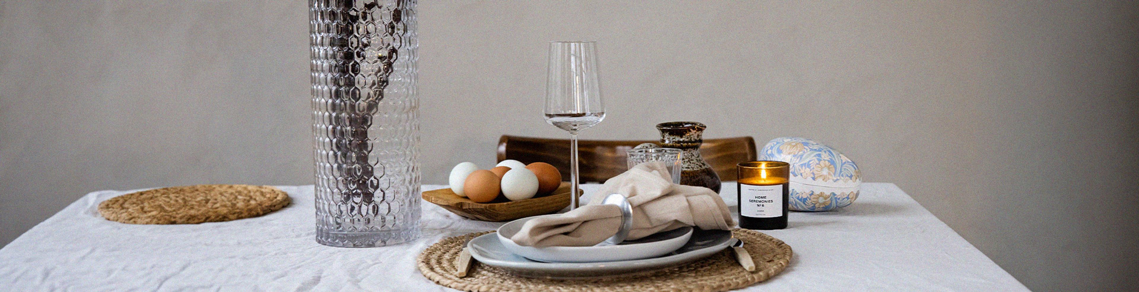 Ett dukat bord med en vit servett i en ring på en ljus tallrik, ett glas, ett blockljus i glas, en skål med vita och bruna ägg samt en vas med kvistar. En vävd rund duklapp ligger under tallriken.