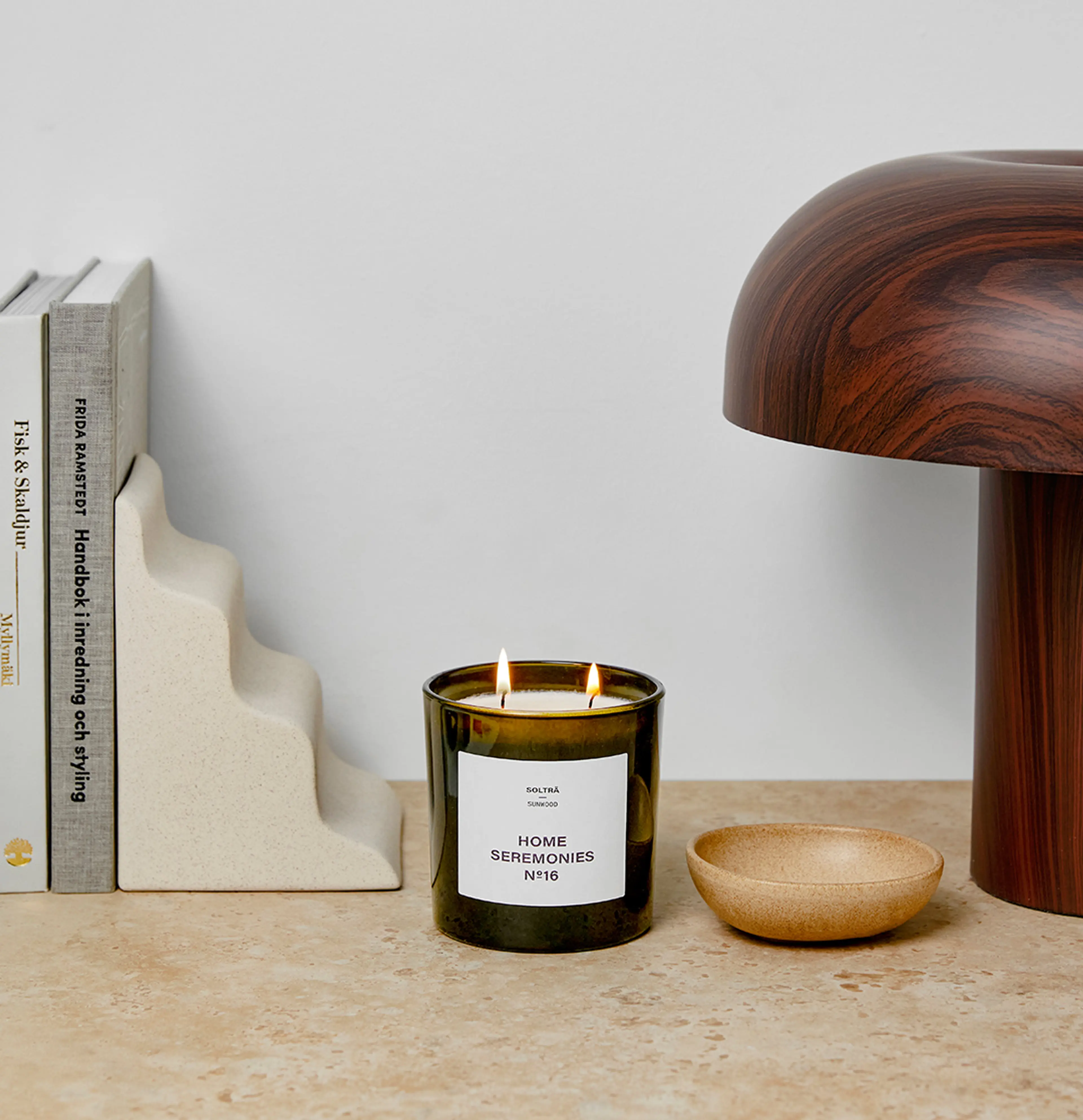 Ett doftljus med två vekar i en mörk glasburk, med vit etikett och texten "Home Seremonies". Ljuset står på en bänk bredvid några böcker, en keramisk bokstöd och en skål i beige ton.
