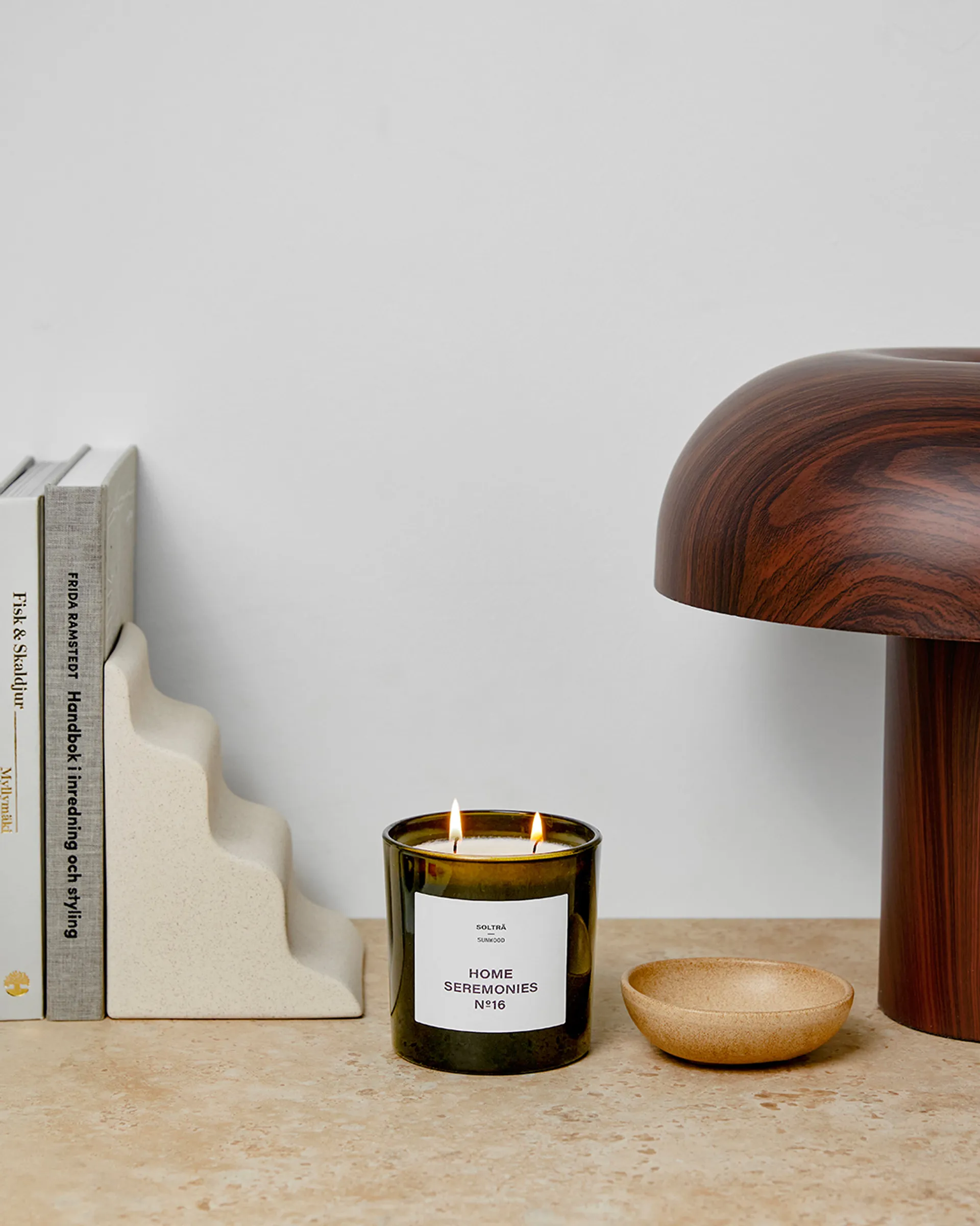 Ett doftljus med två vekar i en mörk glasburk, med vit etikett och texten "Home Seremonies". Ljuset står på en bänk bredvid några böcker, en keramisk bokstöd och en skål i beige ton.