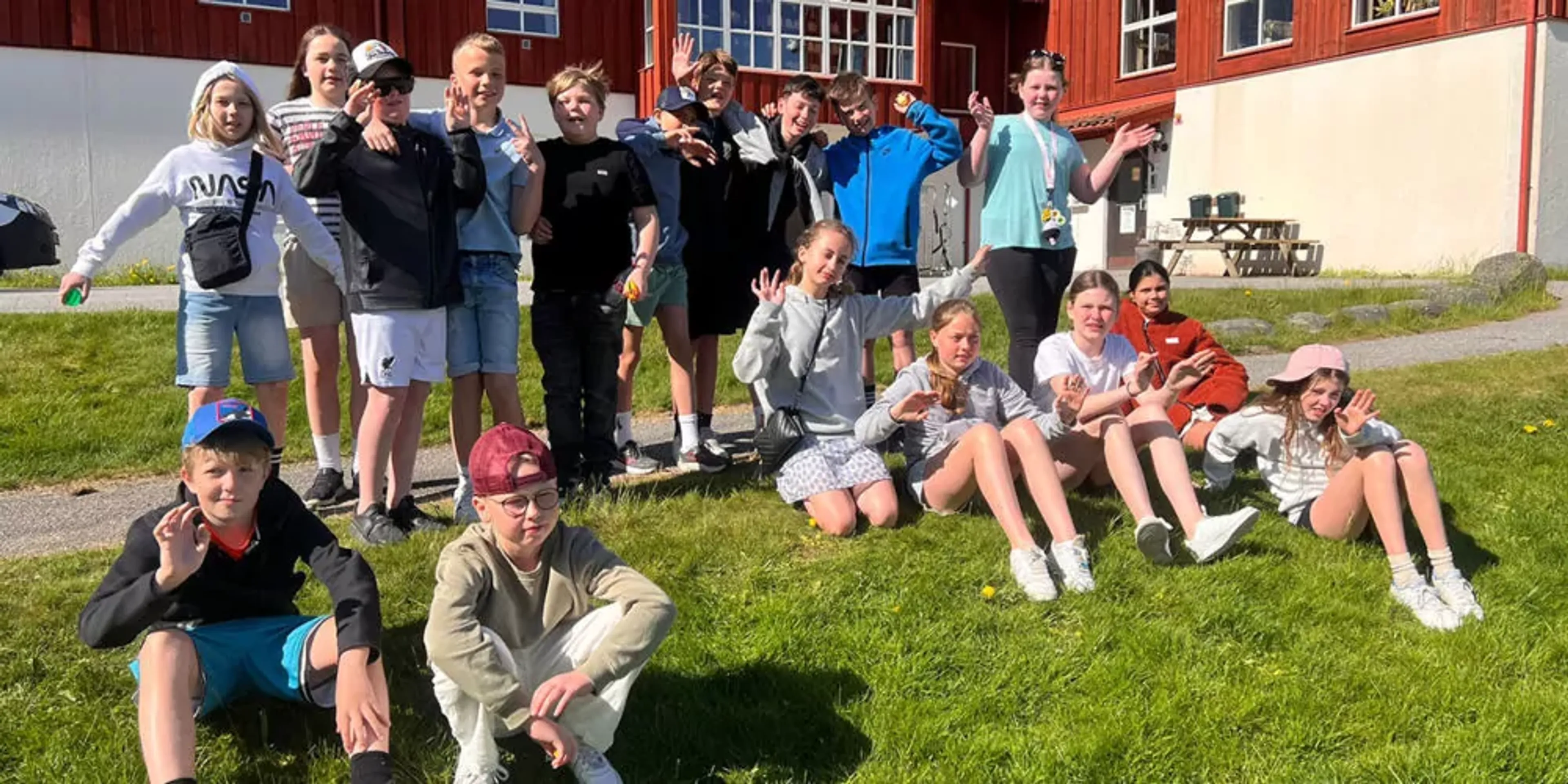 En grupp elever från klass 5B på Hagaskolan samlade utomhus i solsken, en del sitter på gräset medan andra står bakom dem. Bakgrunden visar en skolbyggnad i rött och beige.