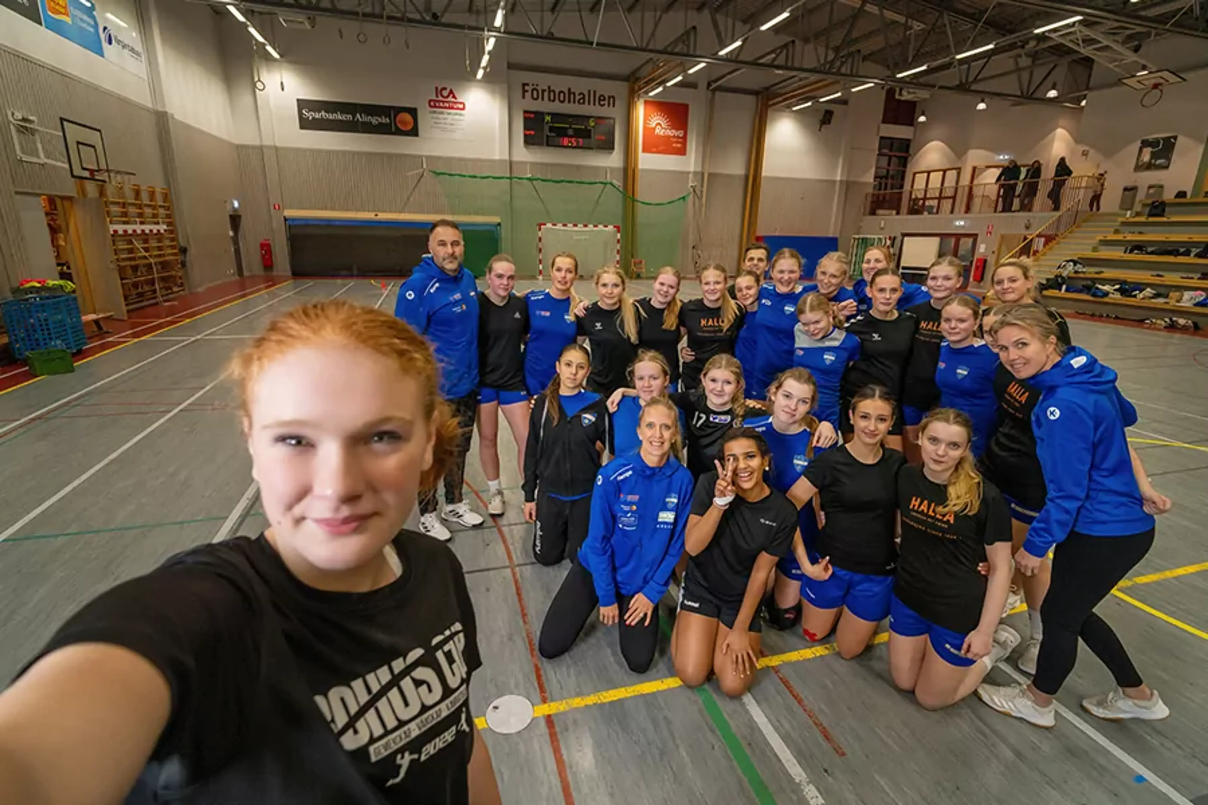 En grupp unga handbollsspelare och tränare samlade i en sporthall, där en av spelarna tar en selfie med hela laget i bakgrunden. Alla är klädda i sportkläder och ser glada ut.