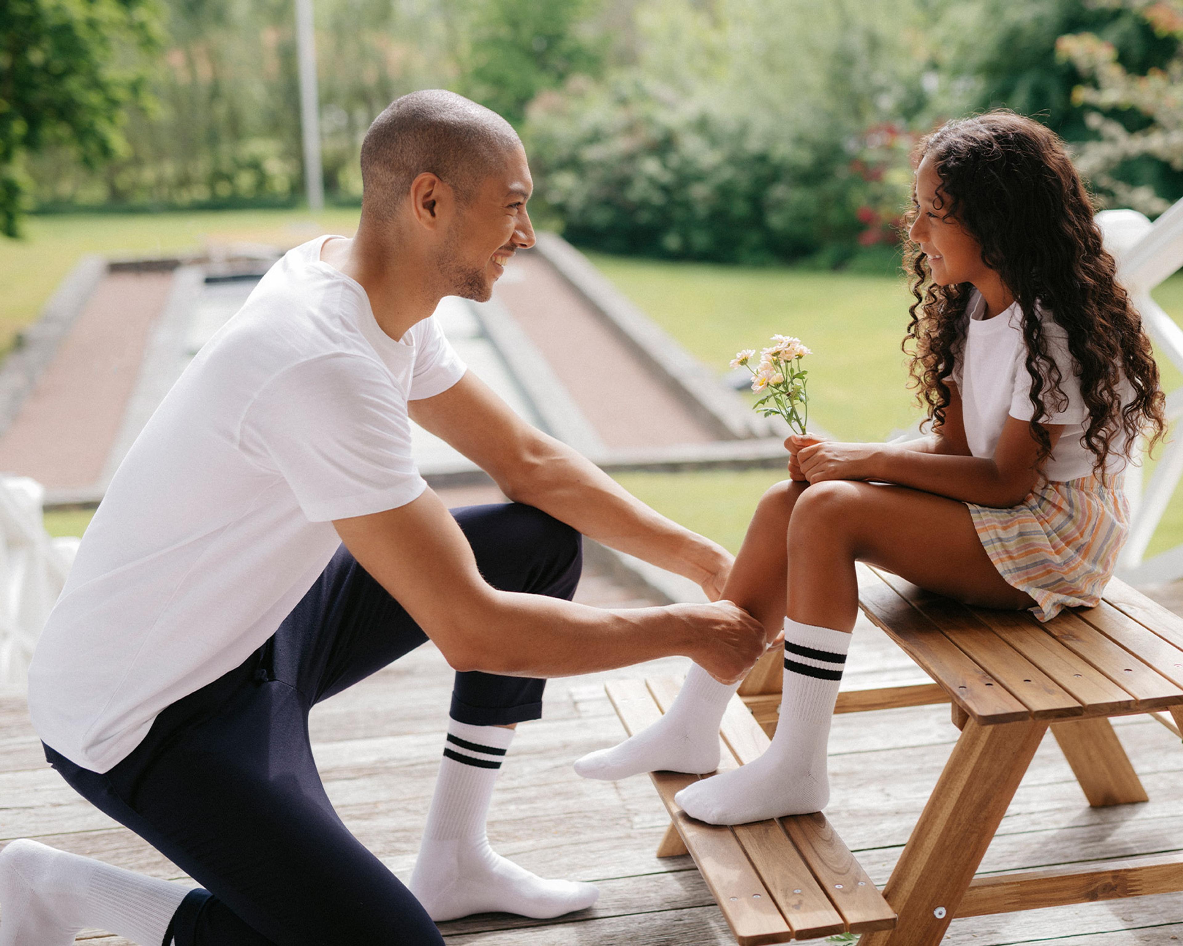 En vuxen man knyter vita sportstrumpor med svarta ränder på en flicka som håller små blommor, utomhus på en träbänk. Båda bär matchande strumpor och har på sig somriga kläder.