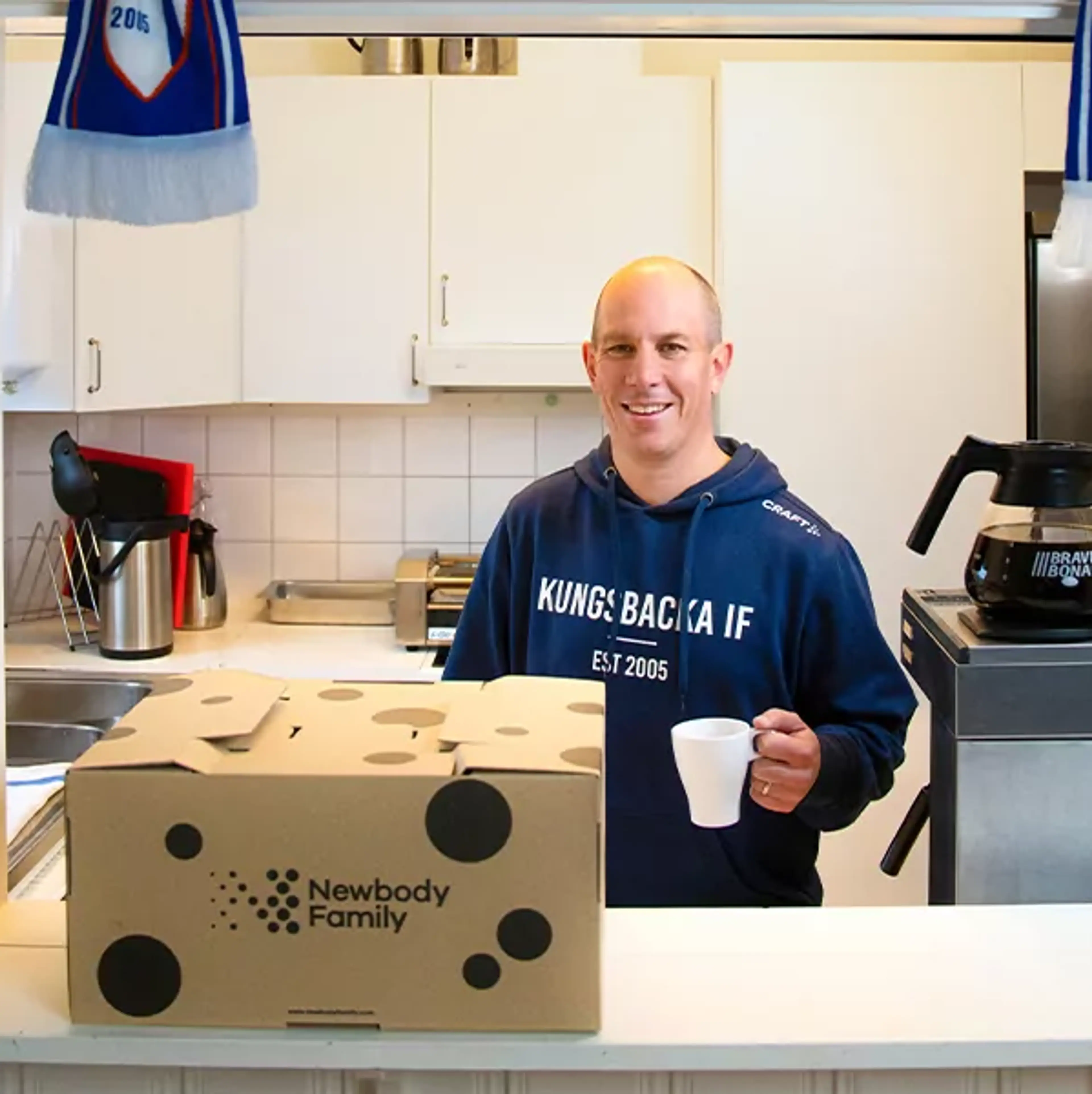 En leende man i en kökslucka håller en vit kaffekopp, framför honom står en kartong med texten Newbody Family. Två blå-vita halsdukar hänger ovanför och visar Kungsbacka IF.