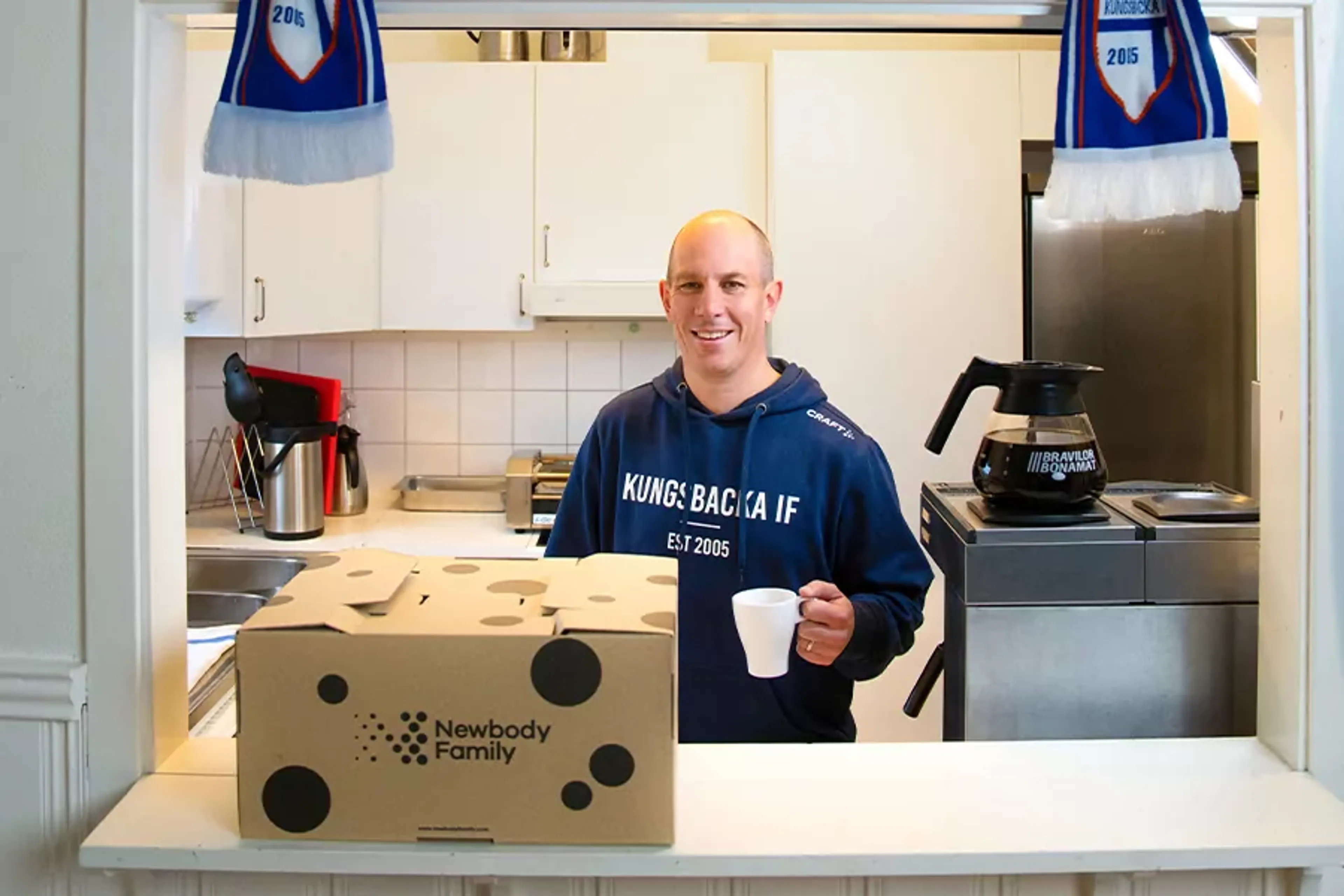 En leende man i en kökslucka håller en vit kaffekopp, framför honom står en kartong med texten Newbody Family. Två blå-vita halsdukar hänger ovanför och visar Kungsbacka IF.