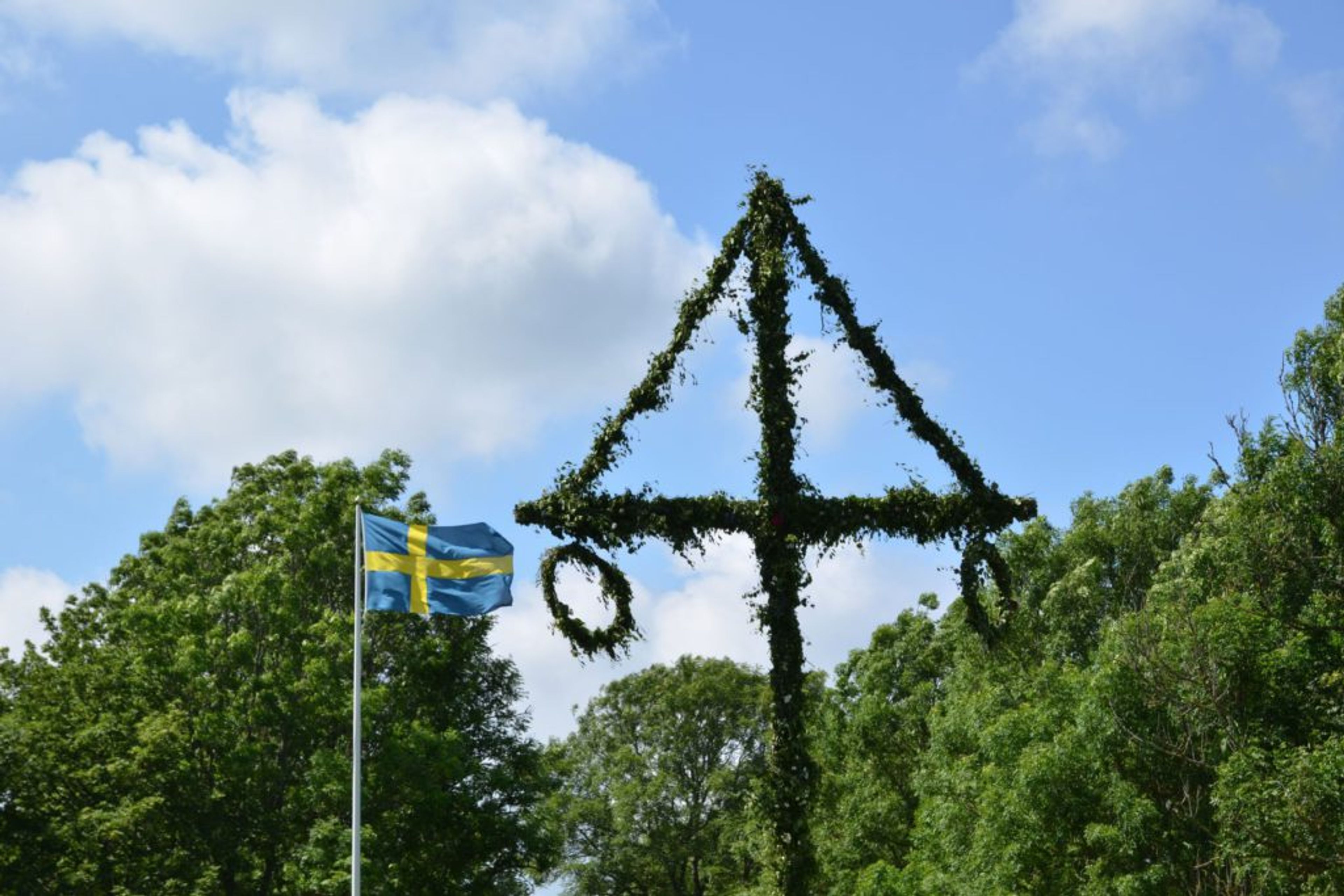 En traditionell midsommarstång dekorerad med gröna kvistar och slingor, omgiven av frodig grönska under en blå himmel. En svensk flagga vajar bredvid i lätt sommarbris.
