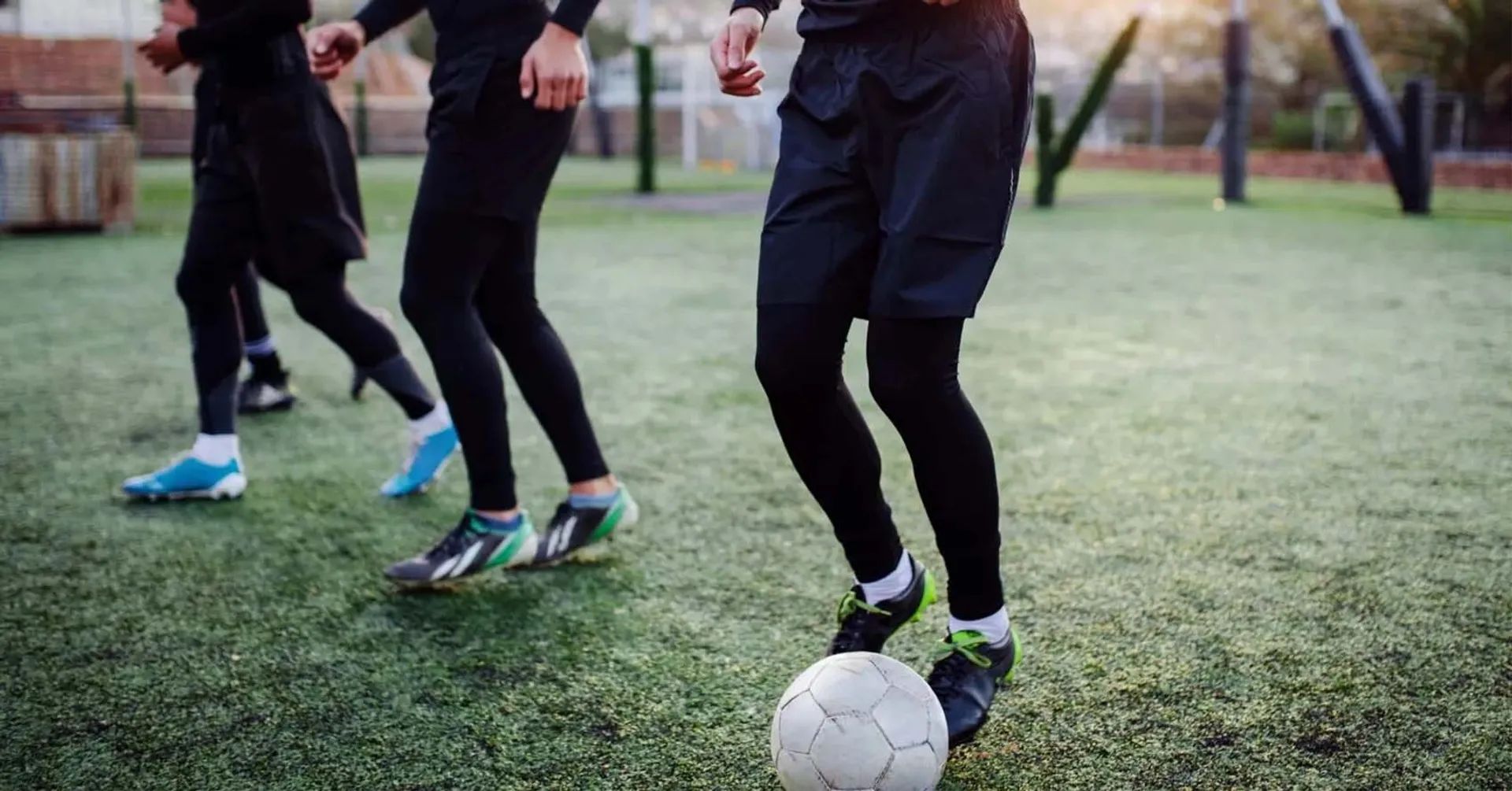 Tre personer tränar fotboll på en grön plan i kvällsljus, alla klädda i svarta träningskläder och sportskor. En vit fotboll syns i förgrunden framför en av spelarna.