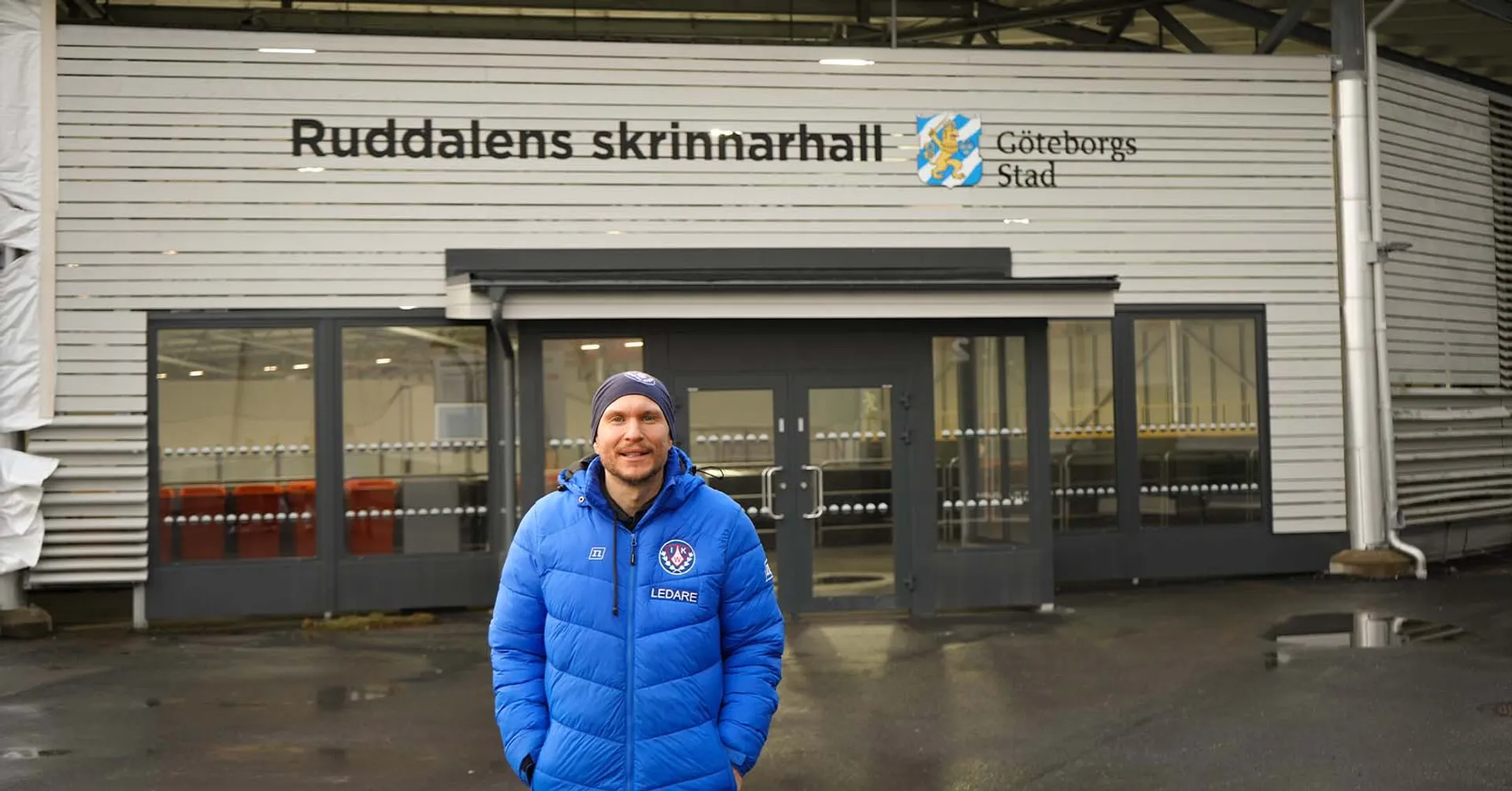 En man i blå jacka med texten "Ledare" står framför entrén till Ruddalens skrinnarhall i Göteborg. Byggnaden är vit och har stora fönster vid ingången.
