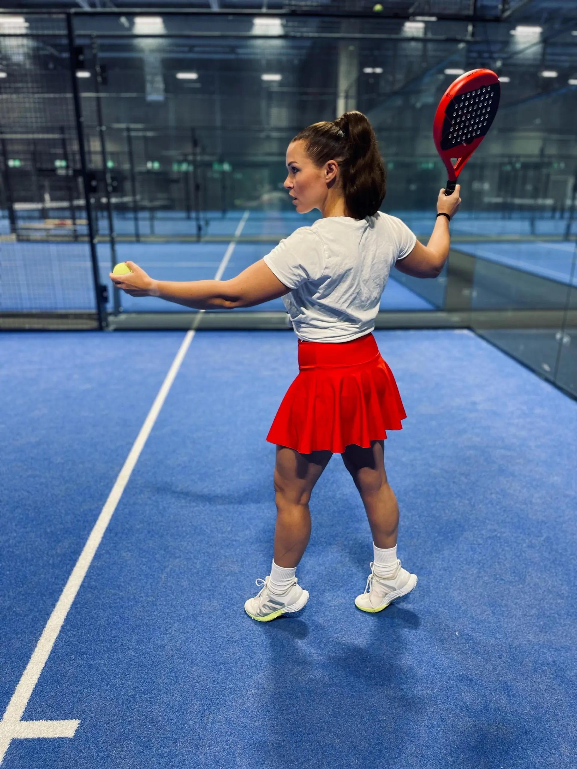 Kvinna i vit t-shirt och röd kjol håller racket redo inför serve på en blå padelbana inomhus. Hon står vänd åt sidan med en boll i handen.